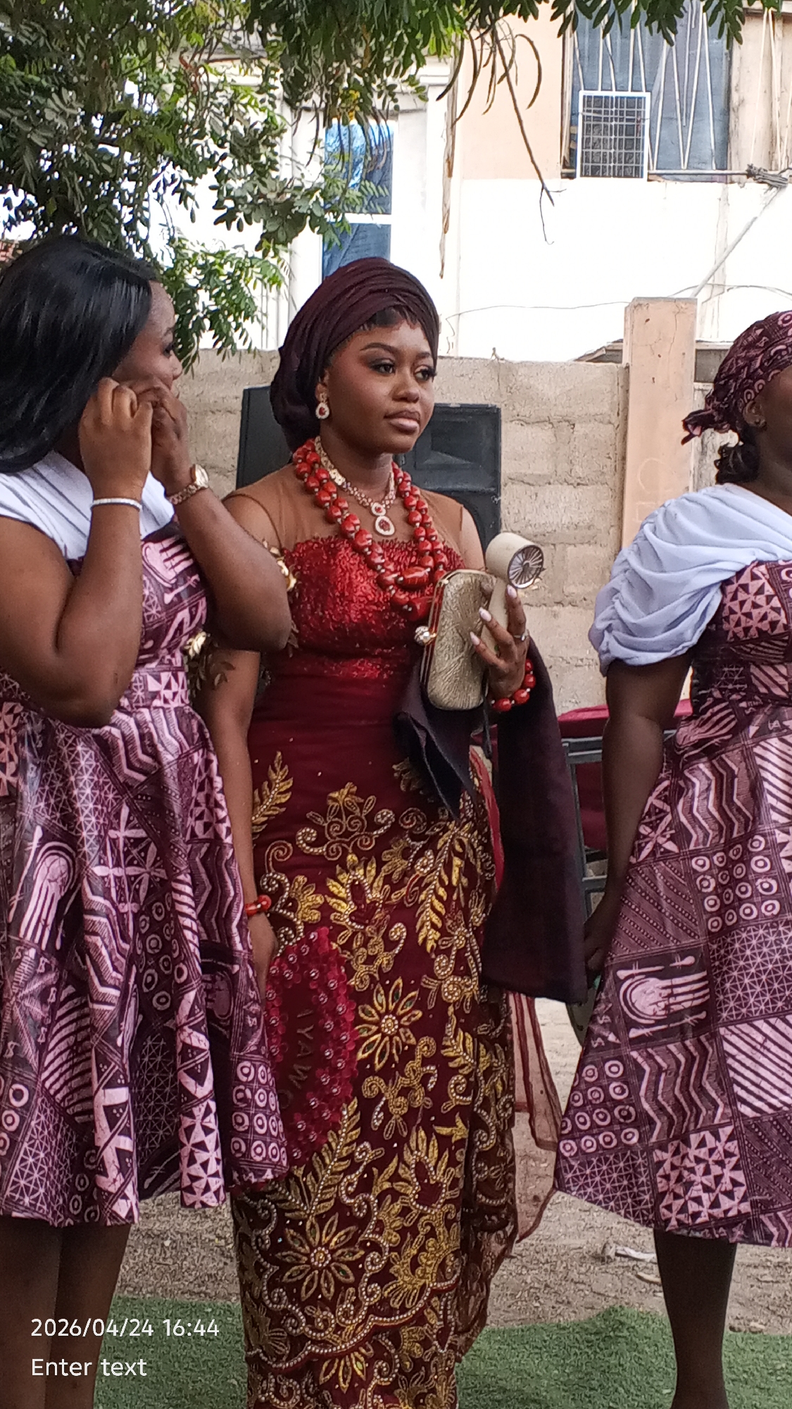 This outdoor, eye-level shot captures three women in traditional attire, likely at a social gathering or celebration during daylight hours.
The central figure is a young woman with dark skin, wearing a dark red headwrap (gele) and an elaborate gown. Her dress features a sequined red bodice with a sheer, nude-colored mesh overlay, and a richly embroidered lower half in red with prominent gold floral and leaf patterns. She is adorned with multiple strands of red coral beads around her neck, matching earrings, and bracelets on her right wrist. She holds a small, ornate gold clutch purse in her left hand and a cylindrical, light-colored object (possibly a portable fan or a small speaker) in her right. Her expression is calm, as she gazes slightly to her right. Visible on the gold embroidery of her dress is the partial word "IYAW".
To her left, a woman is partially visible from the chest upwards. She has long, dark hair and wears a light-colored top over a patterned dress, possibly an ankara print, in shades of purple and white. Her hands are raised near her face, with one hand touching her ear, suggesting she might be adjusting an earring or listening intently.
On the right, another woman is partially visible from the torso downwards, wearing a similar patterned dress as the woman on the left, topped with a light blue or white shawl draped over her right shoulder.
The background shows a simple, light-colored concrete wall or building structure with some windows. Overhead, green foliage and tree branches are visible, indicating an outdoor setting. The ground beneath the women is covered in green grass.
In the bottom left corner of the image, the text "2026/04/24 16:44" and "Enter text" is visible, indicating a timestamp.