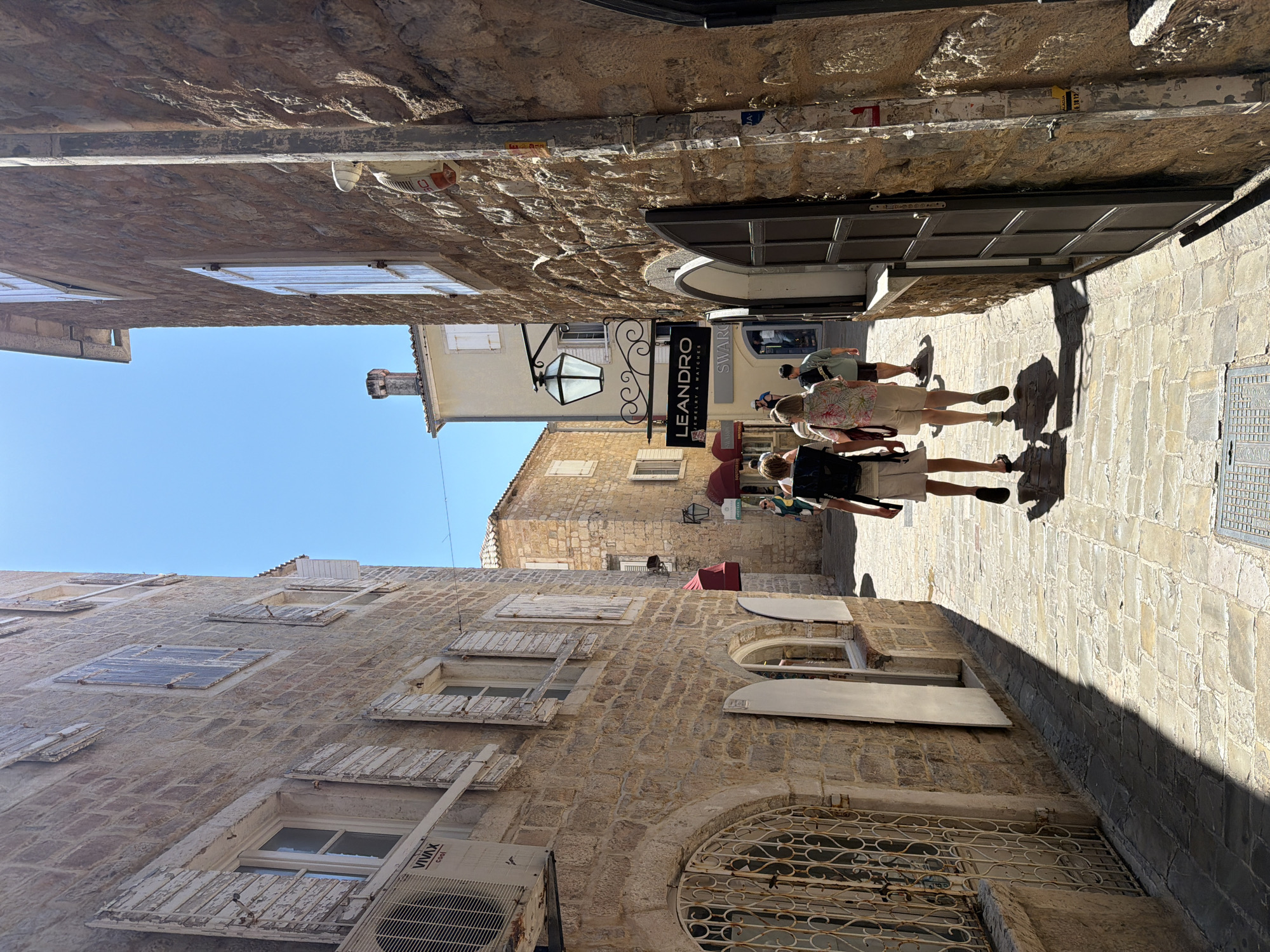 A narrow, cobblestone alleyway in the old town of Budva, Montenegro, is depicted on a sunny day. The alley is flanked by weathered, stone buildings with traditional wooden shutters.
In the foreground, two individuals are walking away from the camera, heading deeper into the alley. The person on the left is wearing a backpack and light-colored shorts, while the person on the right is wearing a floral-patterned shirt and light-colored shorts. Their movements suggest they are tourists exploring the area.
Further down the alley, other people can be seen walking, with some individuals appearing to be looking at shop displays. Signs for businesses such as "LEANDRO JEWELRY & WATCHES" and "SWAROVSKI" are visible, indicating that this is a commercial street. Red awnings extend from some of the buildings, providing shade.
The architecture is characteristic of the Mediterranean, with the stone construction and shuttered windows typical of old European towns. The bright sunlight casts distinct shadows on the cobblestone path, highlighting the textured surfaces of the buildings. The sky is clear and blue, indicating fair weather. An air conditioning unit is visible mounted on the side of one building on the left.