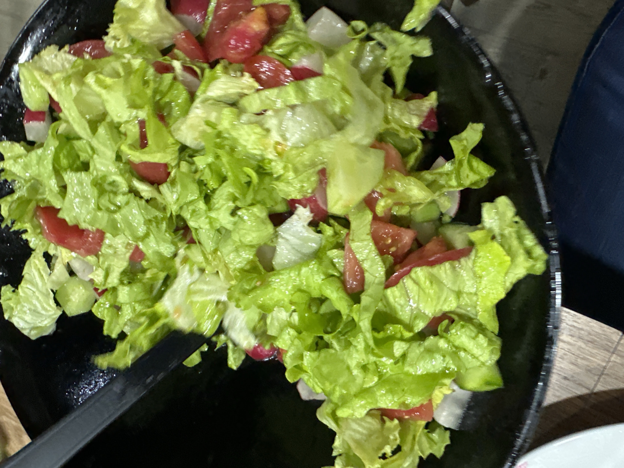 A close-up shot depicts a black bowl filled with a salad. The salad consists of lettuce, tomatoes, and radishes. A black serving utensil is partially visible at the bottom left of the frame. The salad sits on a wooden surface. A portion of a blue garment is visible in the upper right corner. The bottom right of the frame shows a white plate. The location is not discernible from the image.