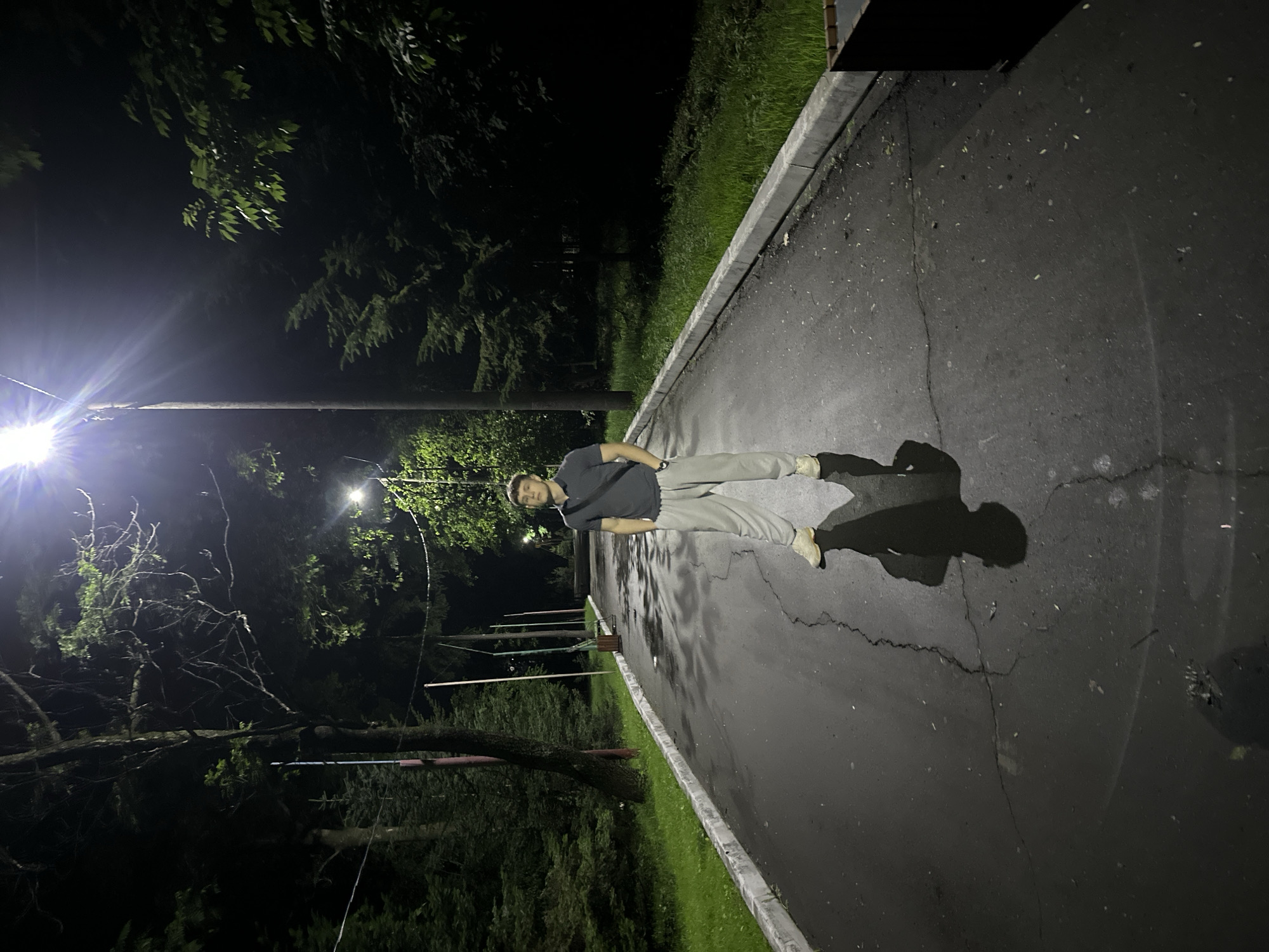 A young man stands in the middle of a wet asphalt path at night, illuminated by a bright street lamp overhead. He is wearing a dark grey polo shirt and light grey sweatpants, with beige sneakers. His hands are in his pockets, and he is looking towards the camera. The path is lined with grass and trees on either side. The environment appears to be a park or a quiet street in the city of Birobidzhan, Russia, as indicated by the specific location. The street lamp casts a strong glare, creating a distinct shadow of the man on the wet ground. In the background, there are more trees and street lights further down the path.