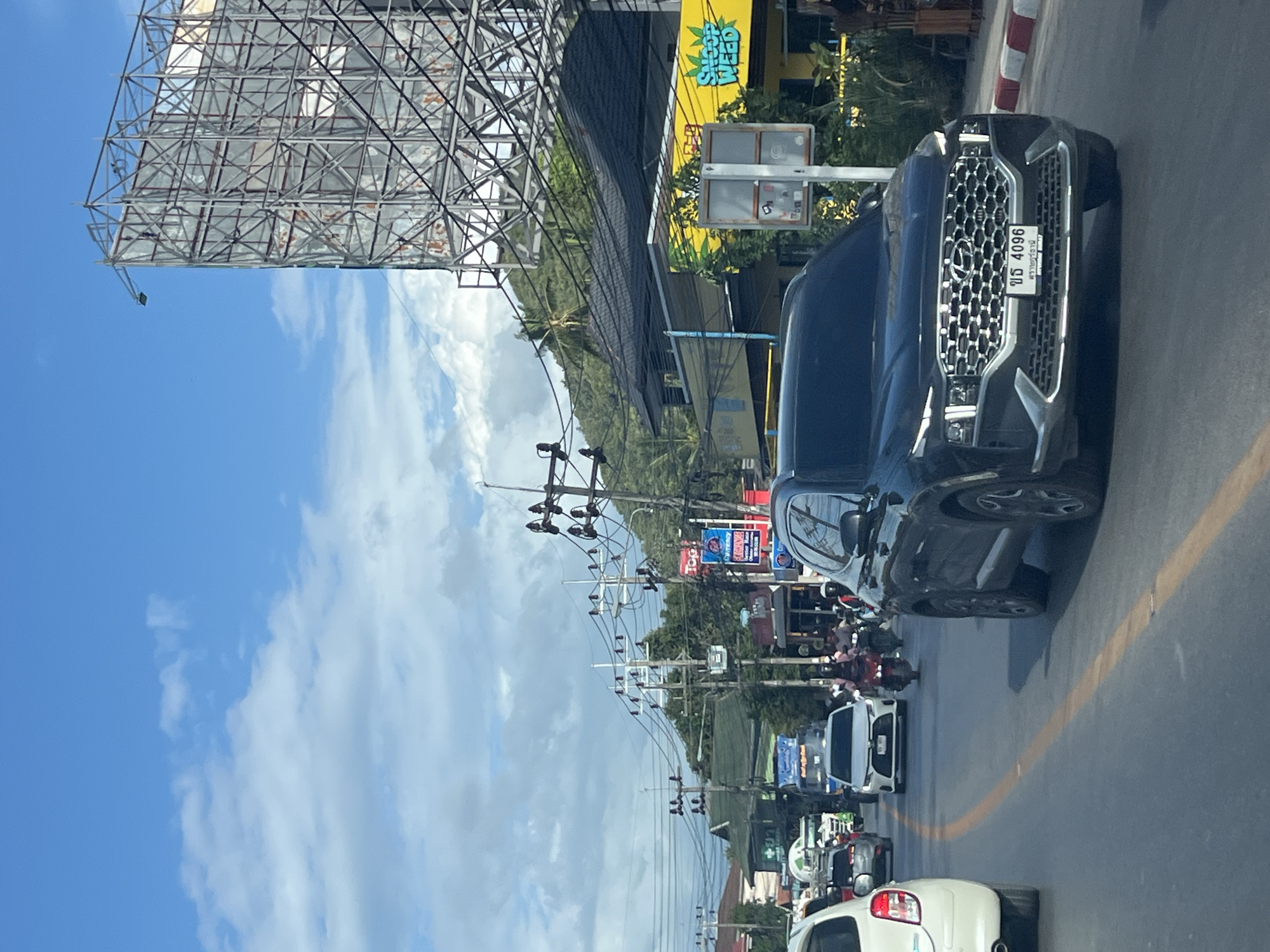 The image depicts a busy street scene in an urban environment. A black Hyundai Santa Fe is in the foreground, driving on the right side of the road. The car's license plate reads "ขร 4096". A white car is partially visible on the left, also in motion. Numerous other vehicles, including cars and motorcycles, are present on the road, indicating moderate traffic.
Along the right side of the street, there are buildings with awnings and signage, including a prominent yellow building with the words "SNOOP WEED" in green lettering. A large, metallic billboard structure stands tall against the blue sky, which is partly cloudy. Trees and lush green hills are visible in the background, suggesting a location with a tropical or subtropical climate. Overhead power lines crisscross the scene. The street is paved and marked with a yellow line. The overall impression is of a commercial area with a mix of businesses and residential structures, experiencing a flow of traffic under a bright sky.