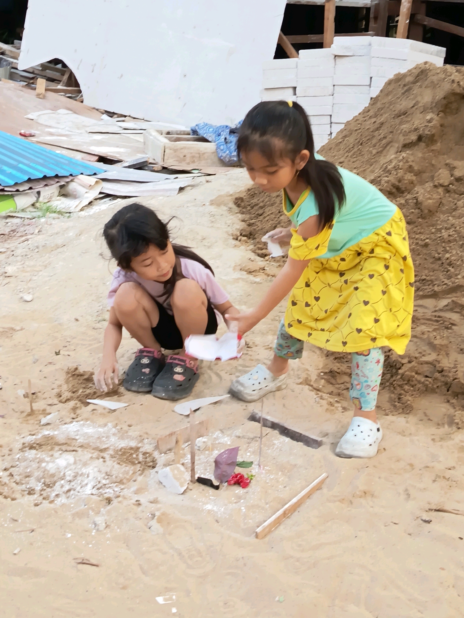 Two young girls are playing in a sandy area, possibly a construction site, in Banjarmasin, Indonesia. One girl, wearing a light purple t-shirt and black shorts, is crouched down, digging or playing with the sand. She is wearing dark-colored clogs with pink accents. The other girl, taller and wearing a teal top and a yellow dress with a heart pattern, is standing and handing a small white object to the first girl. She is wearing light-colored clogs and patterned leggings. The environment is sandy with various construction materials visible in the background, including piles of sand, stacks of white bricks, and pieces of metal sheeting and wood. The lighting suggests it is daytime.