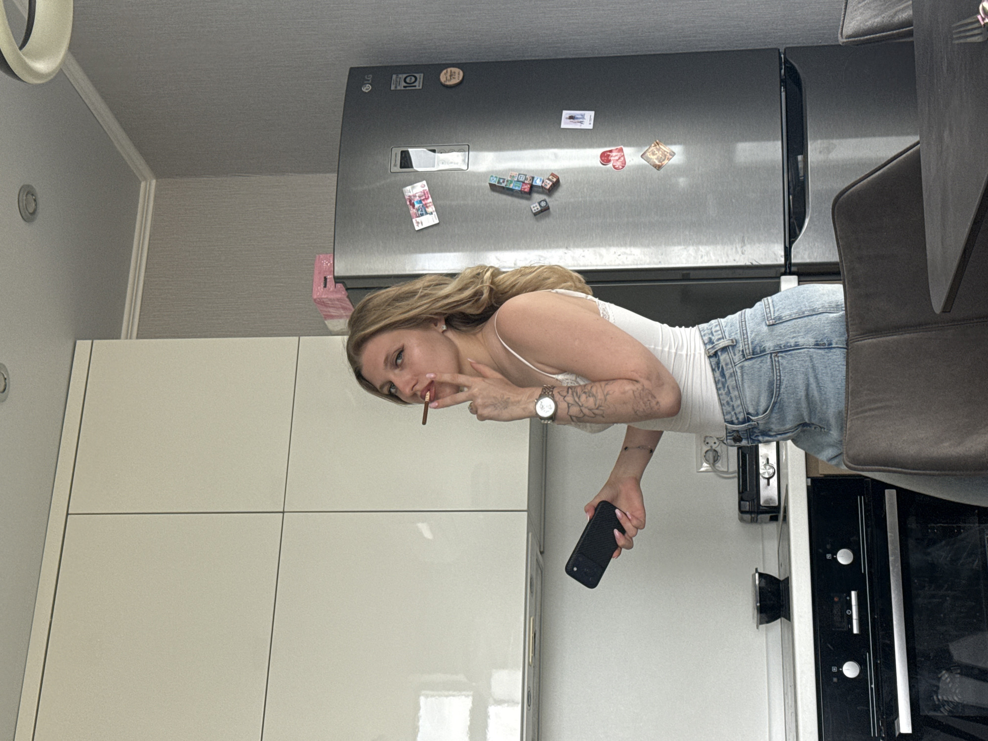 A woman stands in a kitchen. She has blonde hair and wears a white tank top and denim shorts. She is holding a mobile phone in her right hand and appears to be smoking. The kitchen features white cabinets, a stainless steel refrigerator with magnets, and an oven. The location may be Rostov-on-Don, Russia.