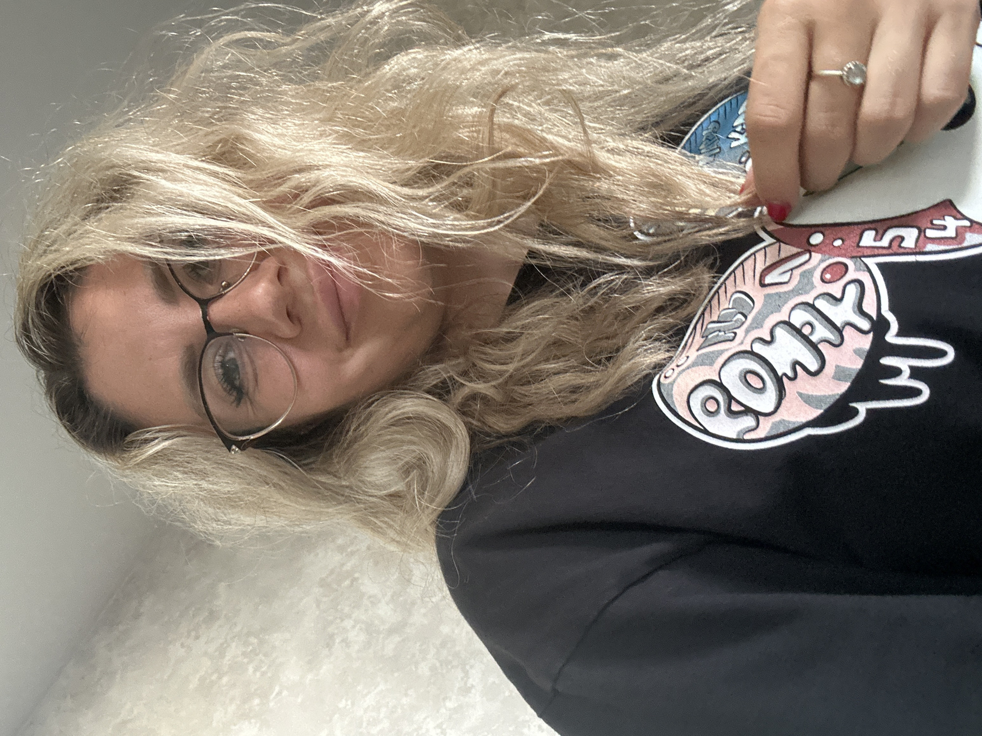 The image is a close-up, selfie-style photograph of a woman with blonde, wavy hair and glasses. She is wearing a black t-shirt with a graphic design on it. Her right hand is visible, holding a white object and wearing a ring. The background appears to be a textured wall. There is no discernible location within the city of Novoaltaysk, Russia, or any other specific geographical information present in the image.
