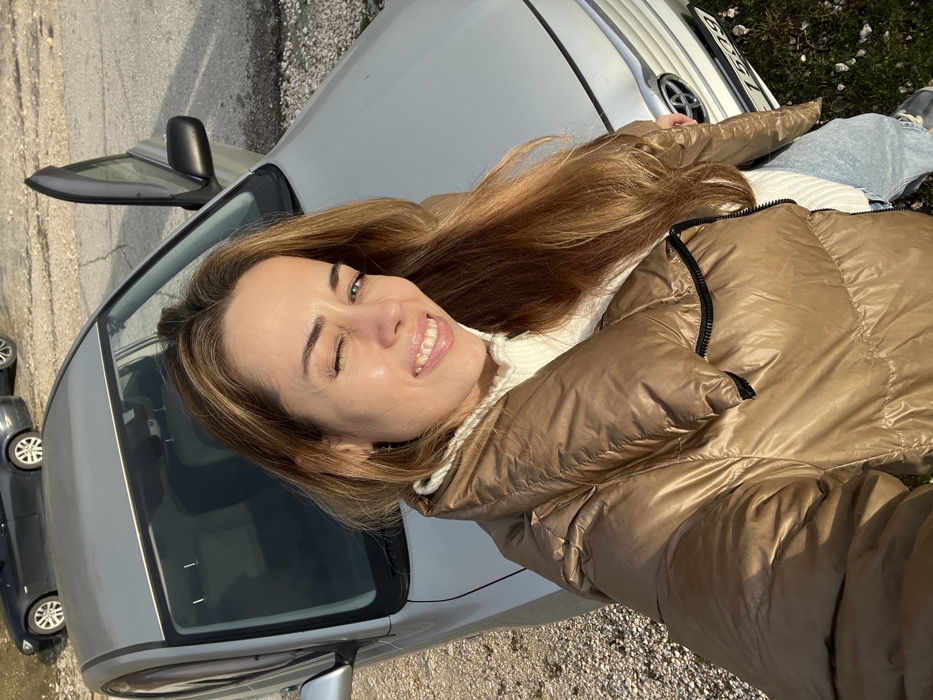 A woman smiles, winking an eye, in a selfie taken next to a silver car. She wears a shiny, golden-brown puffer jacket over a white turtleneck. Light brown hair is visible across her face and shoulder. A car door is open behind her. Another, dark gray car is parked nearby. The background consists of a paved area and some grass. Part of the silver car's license plate is visible.