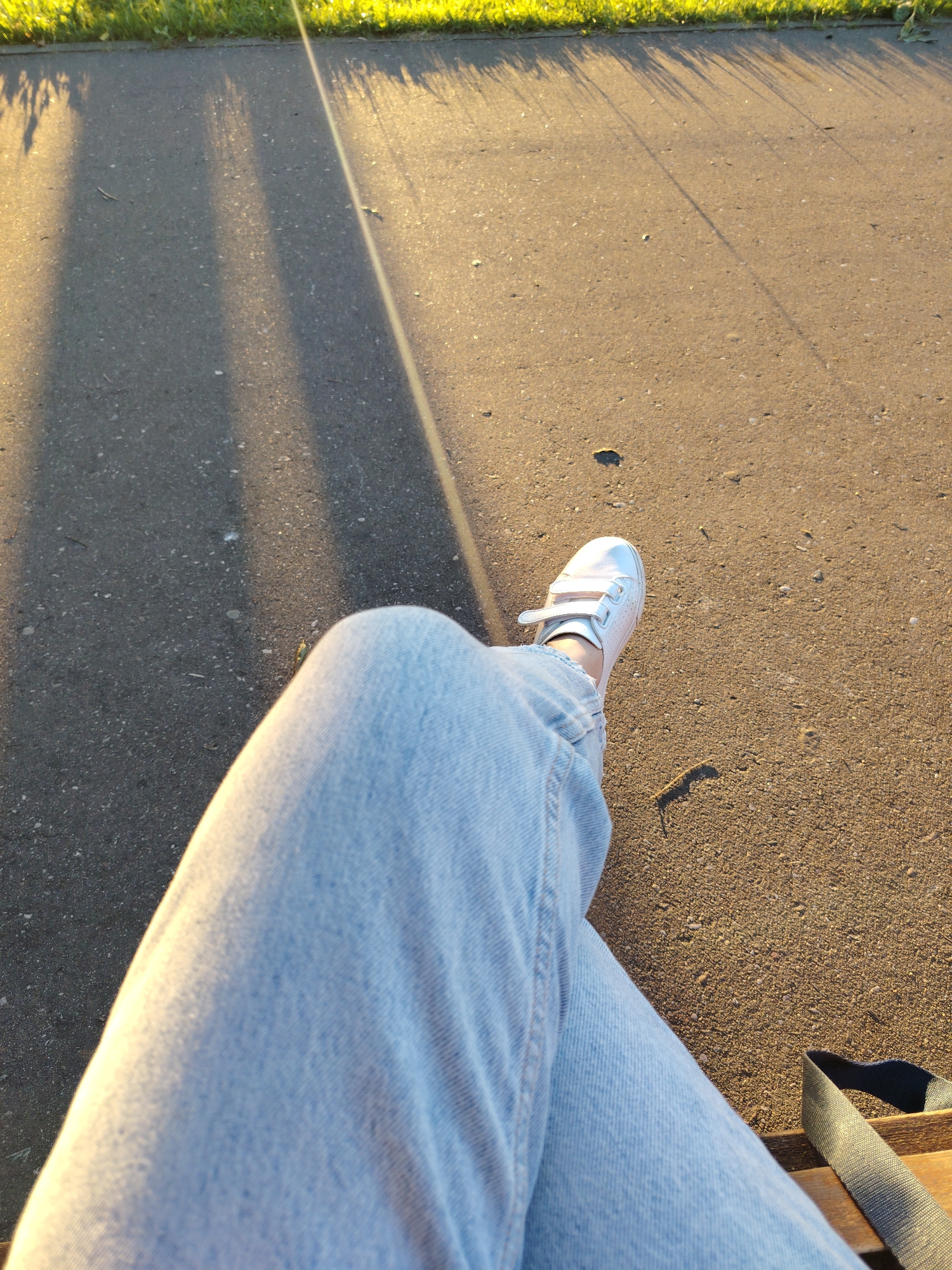 The image shows a person's legs, clad in light blue jeans, resting on what appears to be a wooden bench. One foot, wearing a white sneaker with Velcro straps, is visible. The ground is asphalt, with shadows cast by what seems to be grass or vegetation, illuminated by golden hour sunlight. The environment suggests an outdoor public space, possibly a park or promenade in Konakovo, Russia. A dark strap, likely from a bag, is also visible.