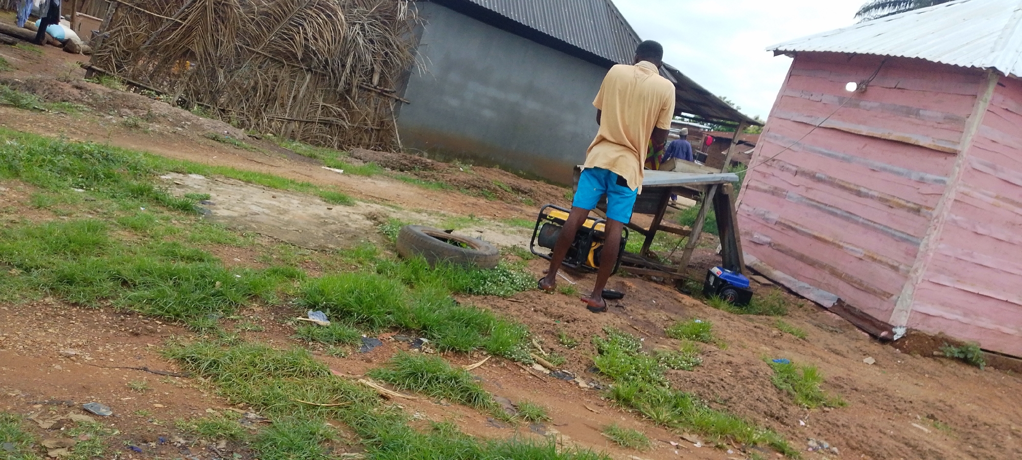 A man in a tan t-shirt and blue shorts stands near a portable generator in a rural or semi-rural setting, possibly a construction site, in Baissa, Nigeria. He is facing away from the camera, looking towards a wooden table. In the background, another person in a blue shirt is visible behind the table. A pink wooden structure with a corrugated metal roof is to the right of the frame, and a larger building with a grey wall and corrugated metal roof is in the mid-ground to the left, with a large stack of dried palm fronds leaning against it. A discarded tire lies in the grass in the foreground, and another smaller generator is on the ground near the pink building. The ground is a mixture of dirt and sparse green grass. The sky is overcast.