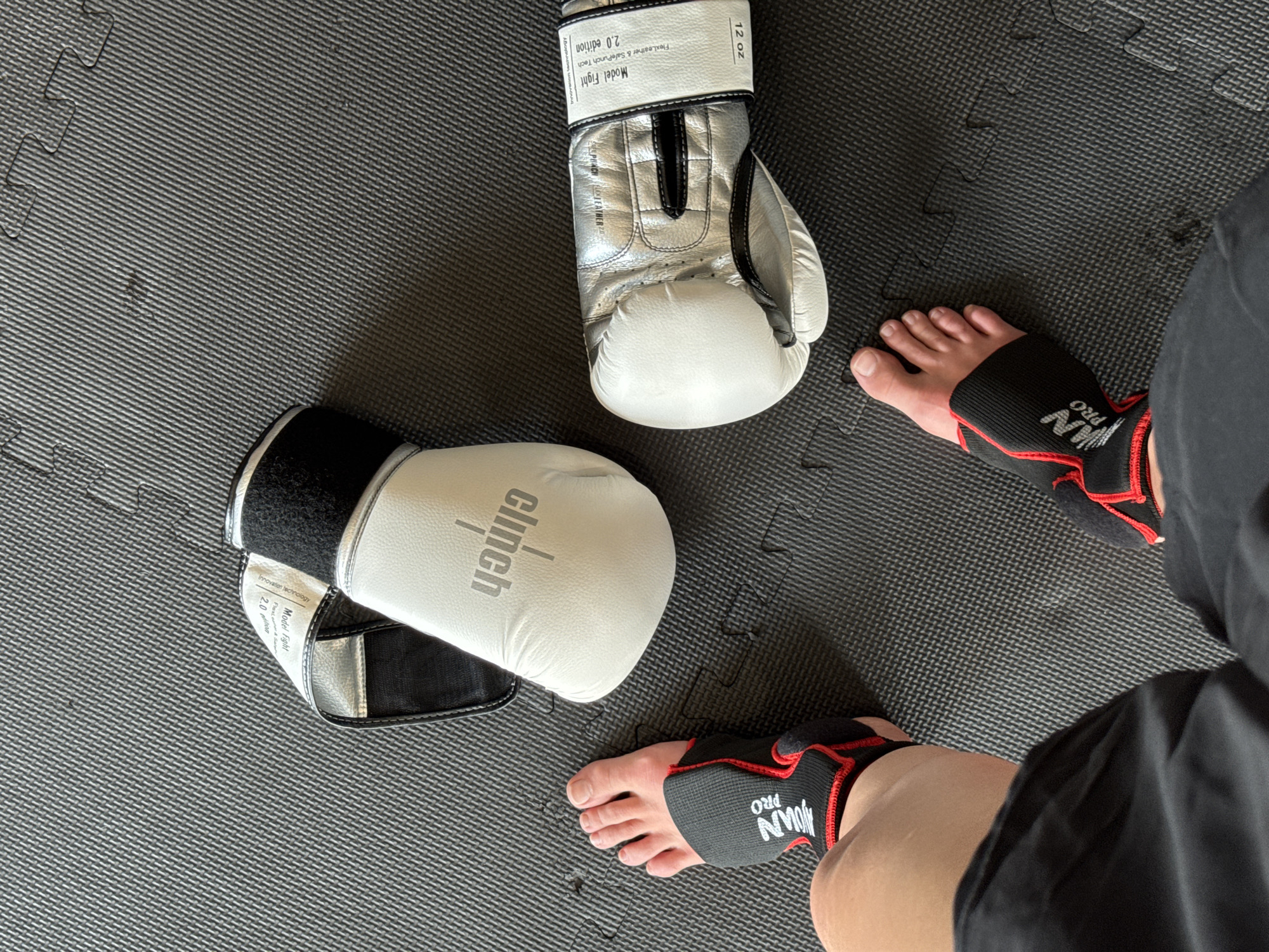 The image is a close-up shot of boxing equipment and a person's feet. The scene appears to be indoors, possibly a gym or training area, based on the interlocking grey floor mats. There are two boxing gloves present, one white with "clinch" written on it, and the other silver and white with "Model Fight" and "12 oz" visible on the wrist strap. A person is visible from the knees down, wearing black ankle supports with red trim, bearing the text "JOUN PRO." The person's toes are visible. There is no indication that this scene is in Tyumen, Russia.