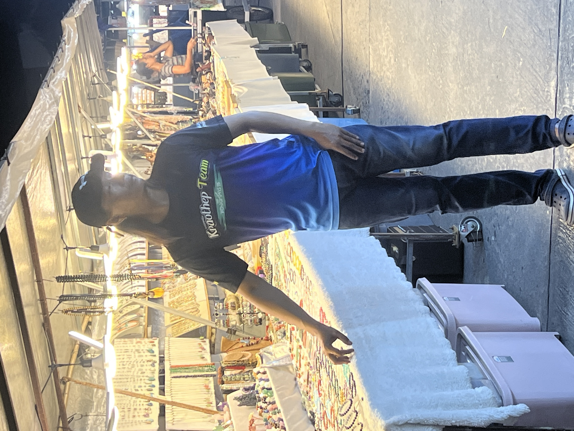 A man, wearing a black baseball cap, a blue and black t-shirt with "Kraothep Team" written on it, and dark jeans, is standing at a market stall. He is looking down and to his left, with his right hand resting on his hip. The stall is filled with various items, including necklaces, bracelets, and other accessories. The market appears to be outdoors, with bright lights illuminating the scene. In the background, other people are visible browsing the stalls, and there are several racks of merchandise. The ground is paved and appears to be a concrete surface. Two pink luggage pieces are visible on the right side of the image. The overall impression is a bustling night market in Thailand, possibly in Koh Phangan, given the location context. The time of day is likely evening or night, due to the artificial lighting.