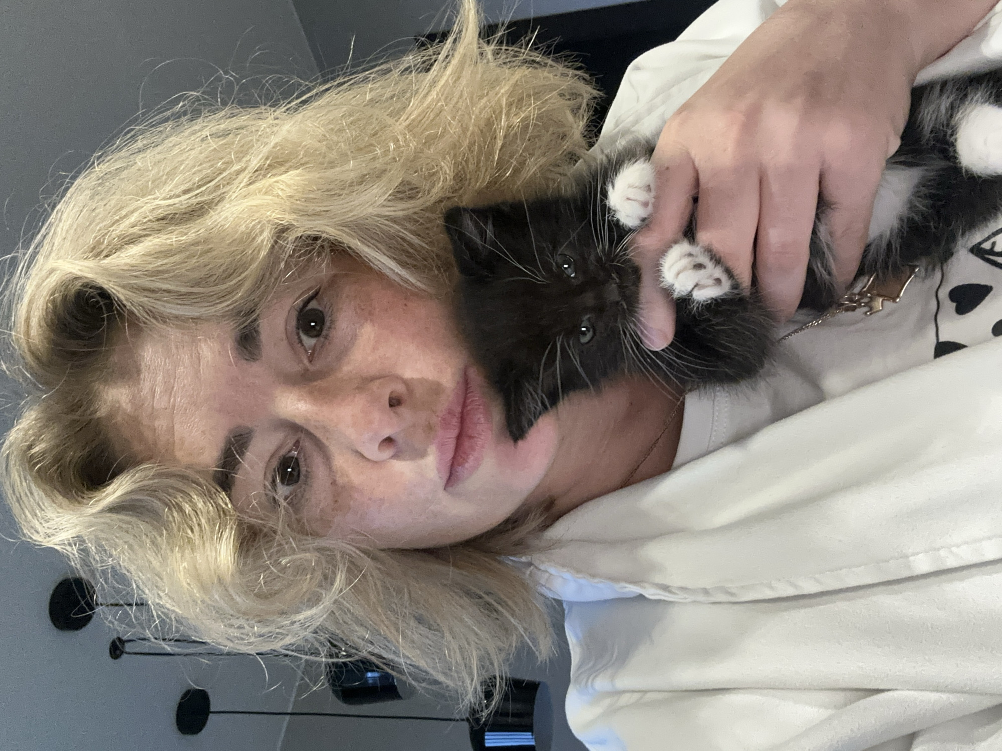 A person with short, wavy blonde hair, wearing a white robe over a t-shirt, holds a black and white kitten. The kitten is small and furry, with white paws and a white chest. The person's face is visible, with light skin and freckles. The background is blurry, but appears to be an indoor setting with lights hanging from the ceiling. The image is taken from a close-up, selfie-style perspective.