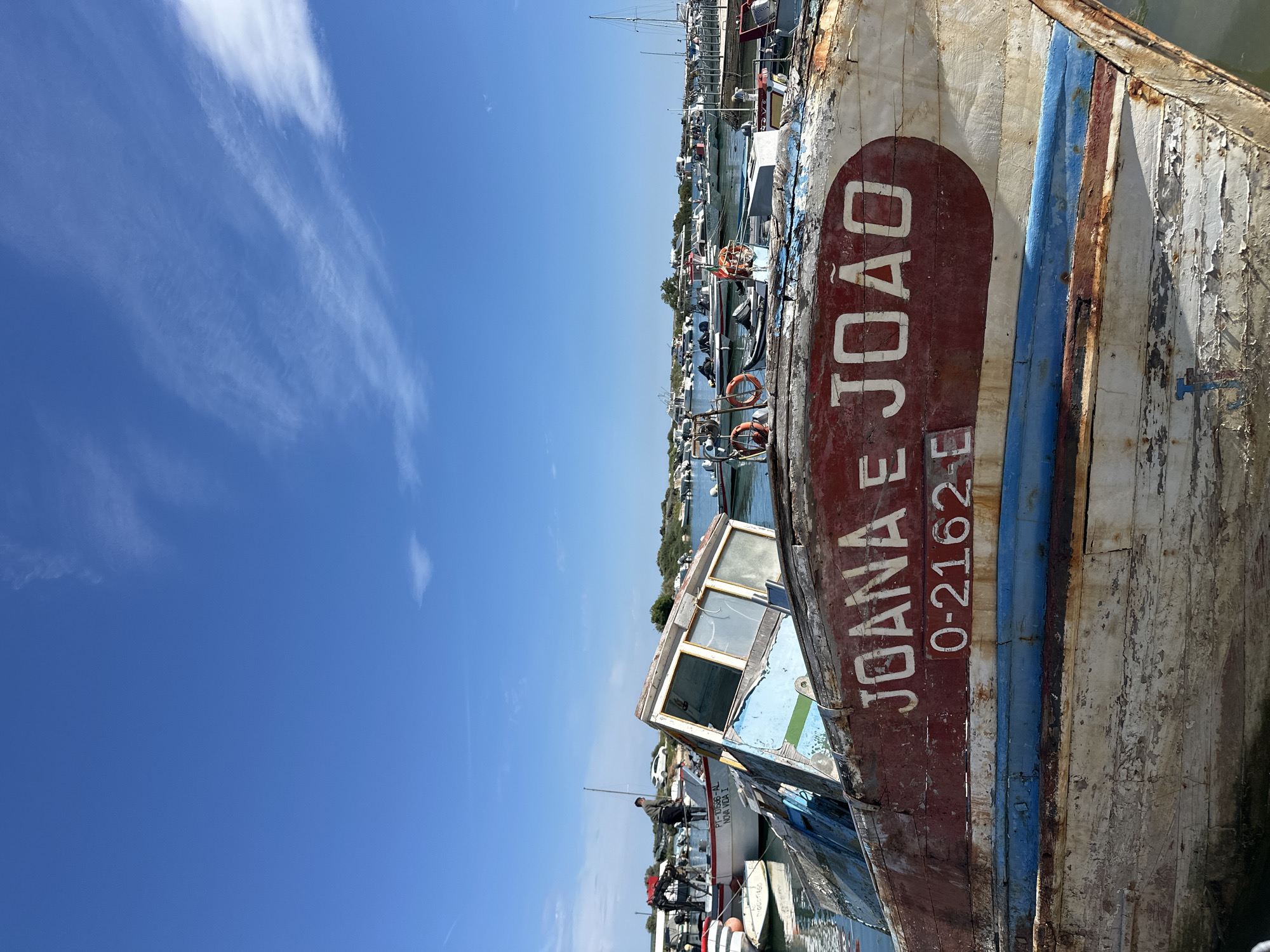 A close-up, rotated image shows a weathered boat named "JOANA E JOÃO" with the identification number "O-2162-E" painted on its hull. The boat's paint is peeling and faded, suggesting age and exposure to the elements. It is docked among other boats in what appears to be a harbor or marina. In the background, more boats are visible, along with buildings and structures along the waterfront. A bright blue sky with scattered wisps of clouds is seen above the boats. The general location is likely in the city of Olhão, Portugal, based on the boat's name and number format.