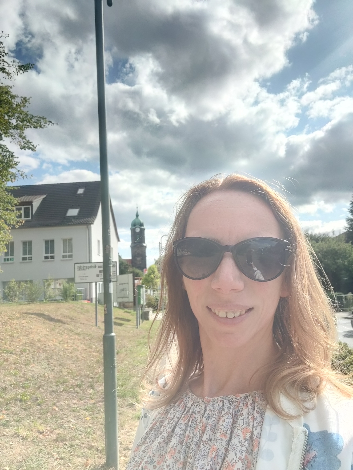 In Mühlsdorf, Germany, a woman with blonde hair and sunglasses takes a selfie outdoors on a cloudy day. She is wearing a floral print blouse and a white jacket. In the background, a two-story building and a clock tower are visible. A lamppost stands in the foreground, obscuring part of the scene. The ground is covered in dry grass.