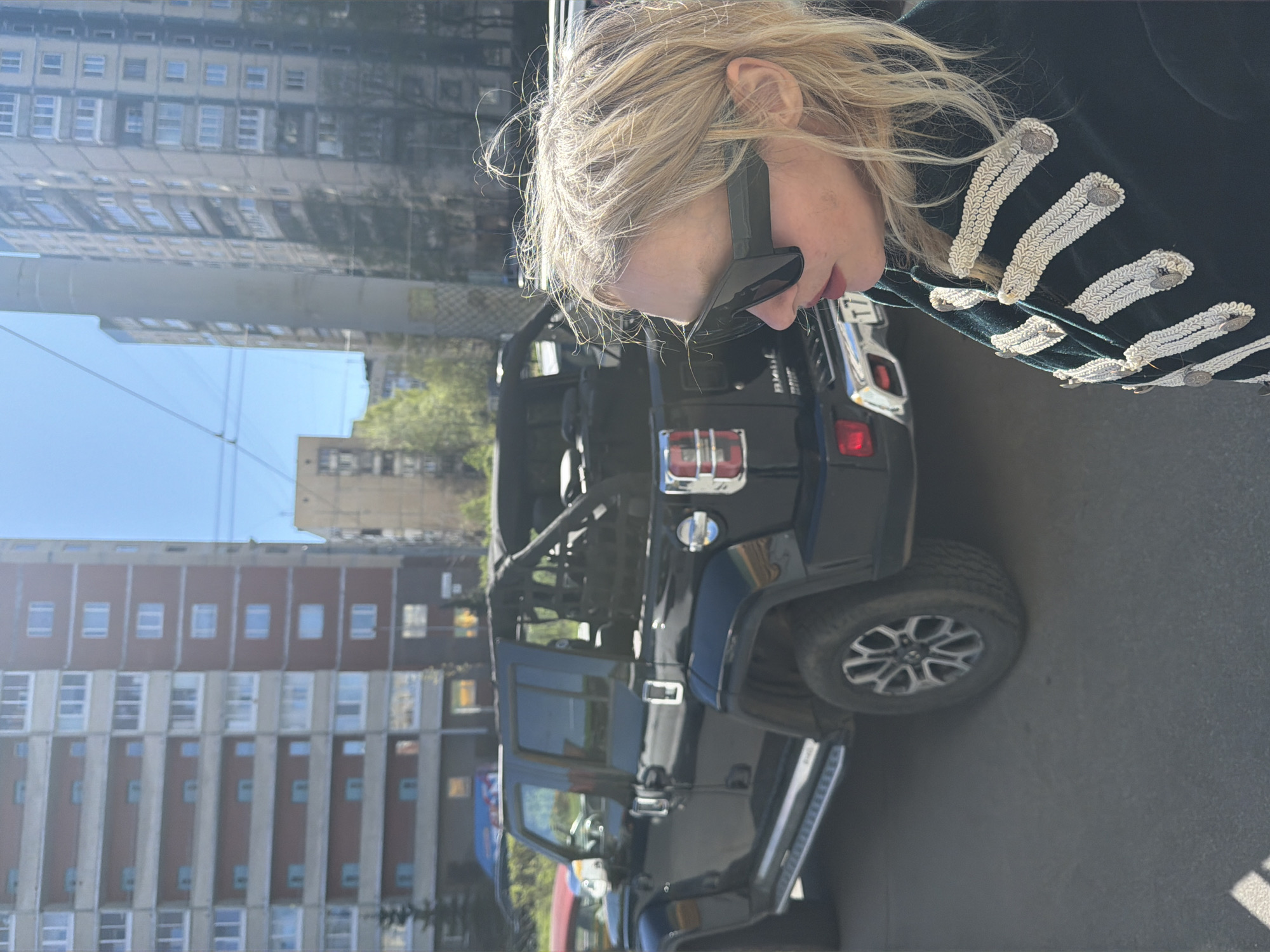 The image shows a fair-skinned blonde woman wearing a black coat with white braided accents on the sleeves, posing with a black Jeep Wrangler in an urban setting. The background features residential buildings, indicating the location may be a city street. Buildings with multiple windows and varying brick colors and heights suggest it could be Saint Petersburg, Russia, known for its diverse architectural styles. The woman wears sunglasses, partially obscuring her face, and her head is turned towards the camera. The vehicle appears to be parked on asphalt, with additional buildings reflected in the car's surface.