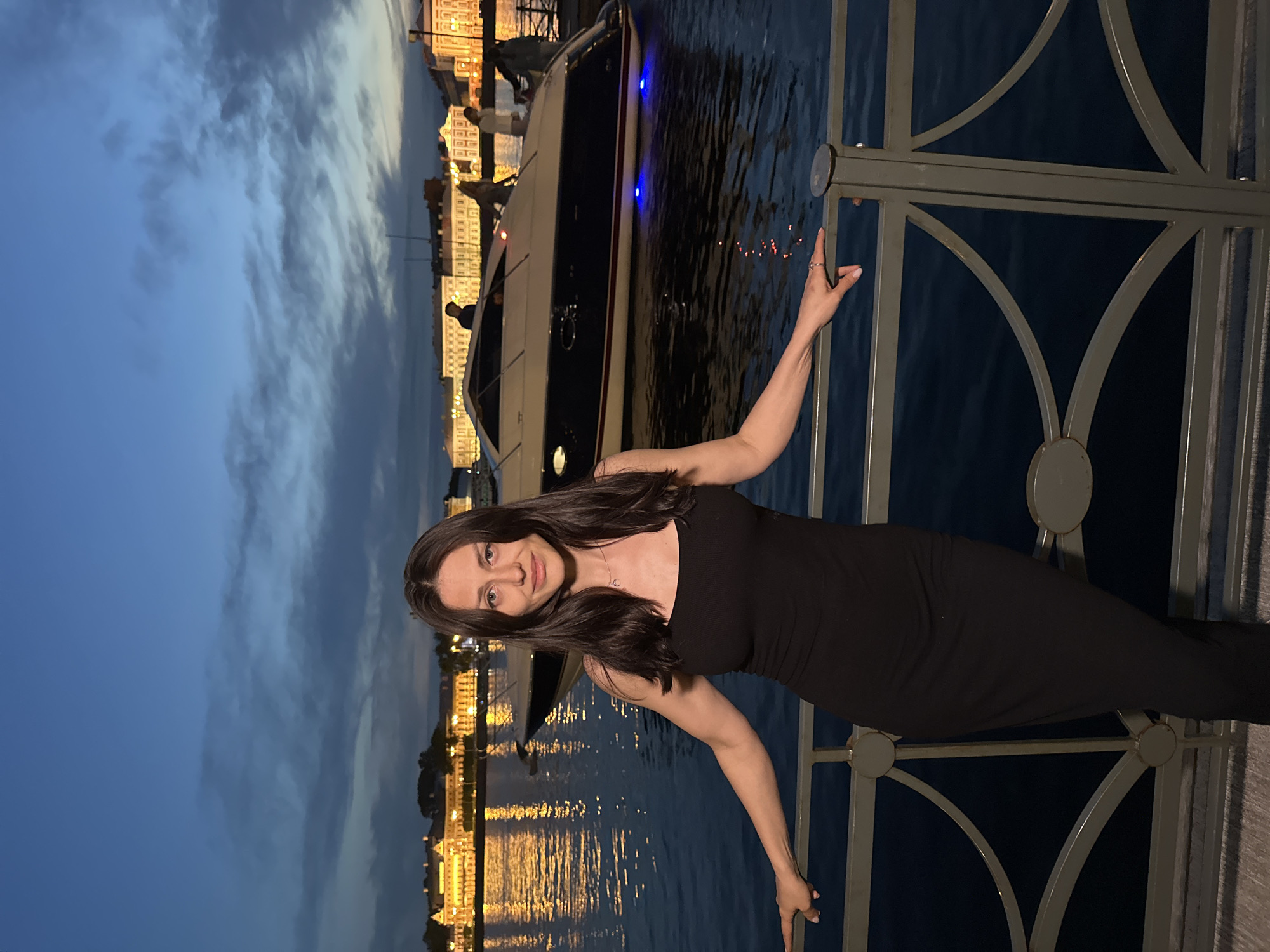 A woman stands on a walkway next to a metal railing, posing for a photo. She has dark hair and is wearing a black dress. Behind her, a body of water reflects lights from buildings and a yacht. The yacht, with blue lights underneath, is moored in the water. In the background, illuminated buildings line the waterfront, suggesting a city location. The sky is a dark blue, indicating it is dusk or night. This scene is likely on the Neva River embankment in Saint Petersburg, Russia, given the architectural style of the buildings and the presence of a river with boats and illuminated embankments.