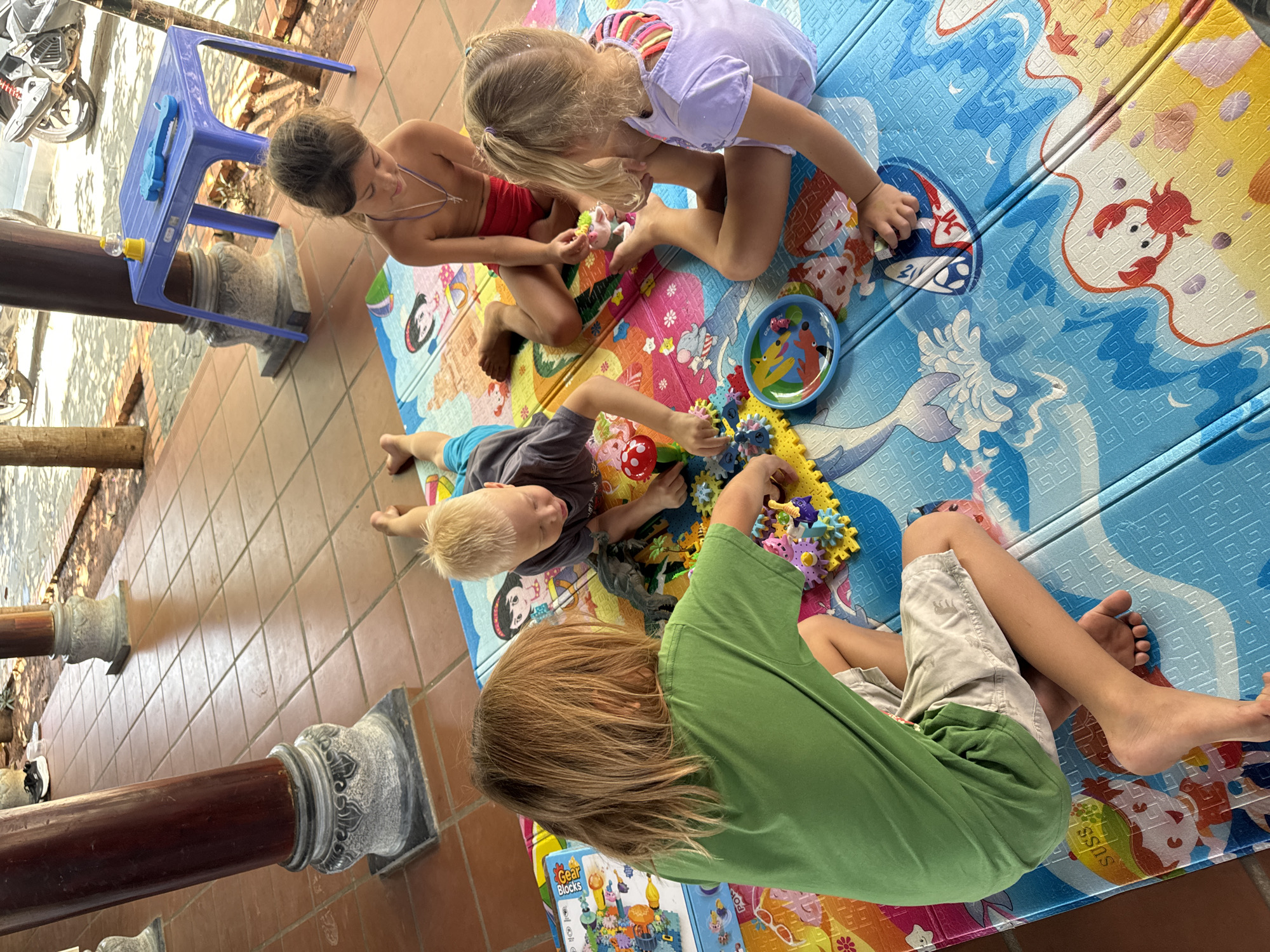 Four children are seen playing together on a colorful, patterned mat laid out on a tiled floor. The scene appears to be an outdoor or semi-outdoor covered area, possibly a patio or veranda, consistent with the location context of Mũi Né, Vietnam, given the terracotta-colored tiles and decorative pillars. Bright natural light illuminates the scene, suggesting it is daytime.
The children are all engaged in playing with various toys spread across the mat. On the left, a girl in a red top with her hair tied back sits attentively. Next to her, another girl in a light purple top with colorful straps, her blonde hair in a ponytail, leans forward, focusing intently on the toys. A boy with blonde hair lies on his stomach, actively manipulating colorful plastic gears. Furthest to the right, another boy with longer brown hair sits with his back to the viewer, also focused on the play.
Among the toys visible are numerous colorful interlocking plastic gears, some partially assembled, a small blue bowl containing miniature figures, a red and white polka-dotted mushroom toy, and a small unicorn plushie. A box labeled "Gear Blocks" is also visible near the boy in the green shirt. The playmat features a variety of cartoon illustrations, including crabs, fish, and other characters, along with abstract designs. In the background to the left, a blue plastic table holds a blue toy, and a silver scooter is partially visible. The children seem calm and engrossed in their activity.