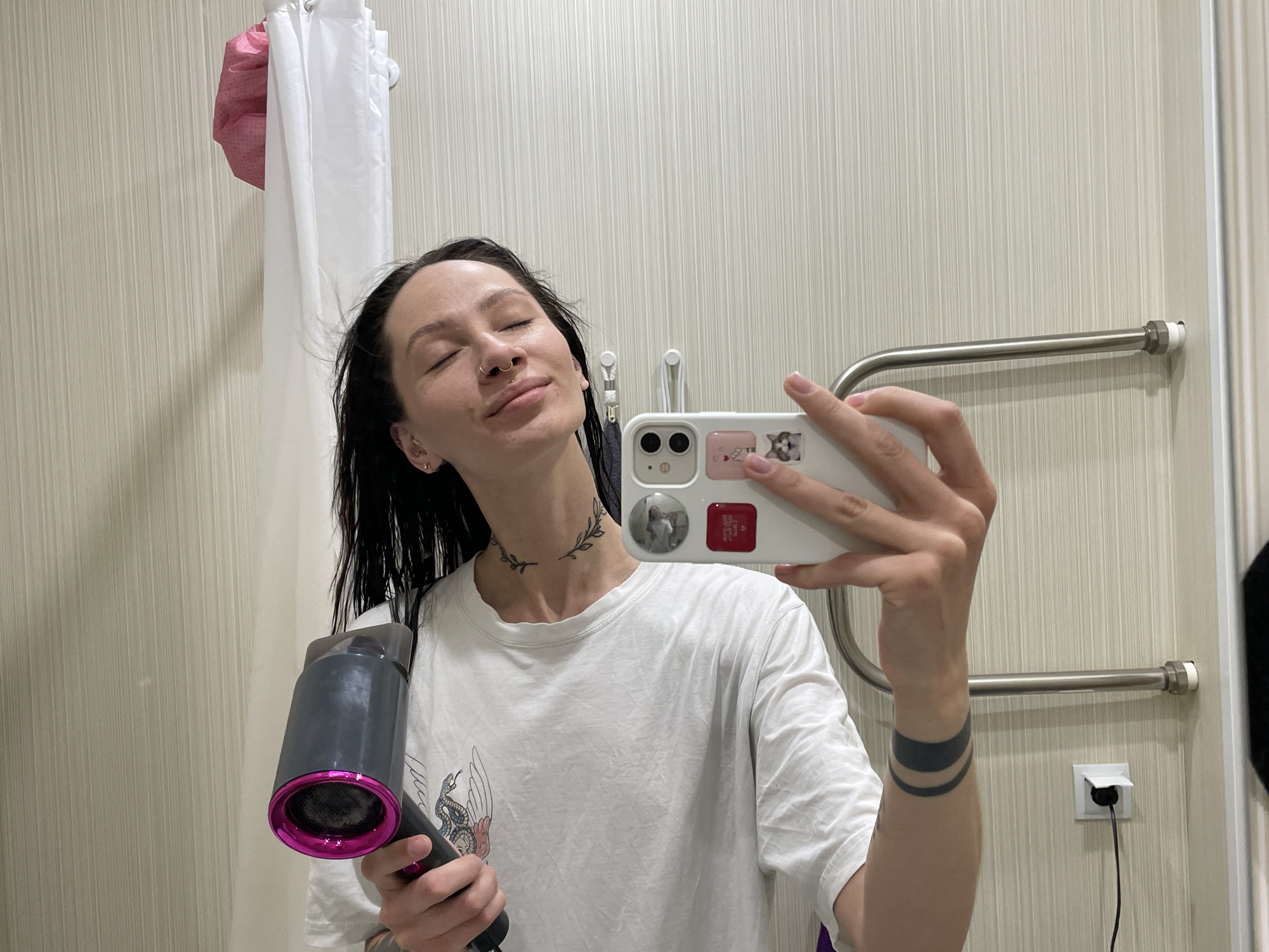 A woman is taking a selfie in a bathroom, holding a hair dryer in her left hand and a phone in her right hand. The bathroom has a beige-colored wall with vertical lines, a heated towel rack, a white electrical outlet with a black cord plugged in, and a partially visible door frame. A pink and white shower cap hangs on a rod next to a white shower curtain. The woman has dark hair and is wearing a white t-shirt. Tattoos are visible on her neck and forearm. The phone has stickers on the back. The location is potentially Izhevsk, Russia.