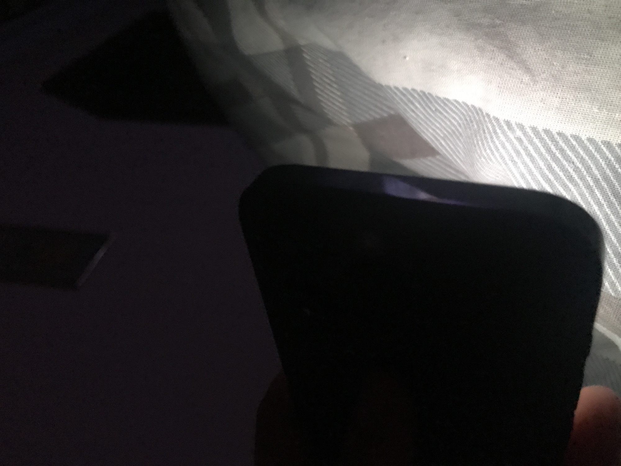 The image shows a dark smartphone positioned against a light-colored, striped fabric. The phone is mostly black and its screen is not illuminated. A hand is visible in the lower right corner of the image. To the left of the phone, in the darkened area, is a small rectangular object. The environment appears to be indoors. The location in the city of Yerevan, Armenia is unknown.