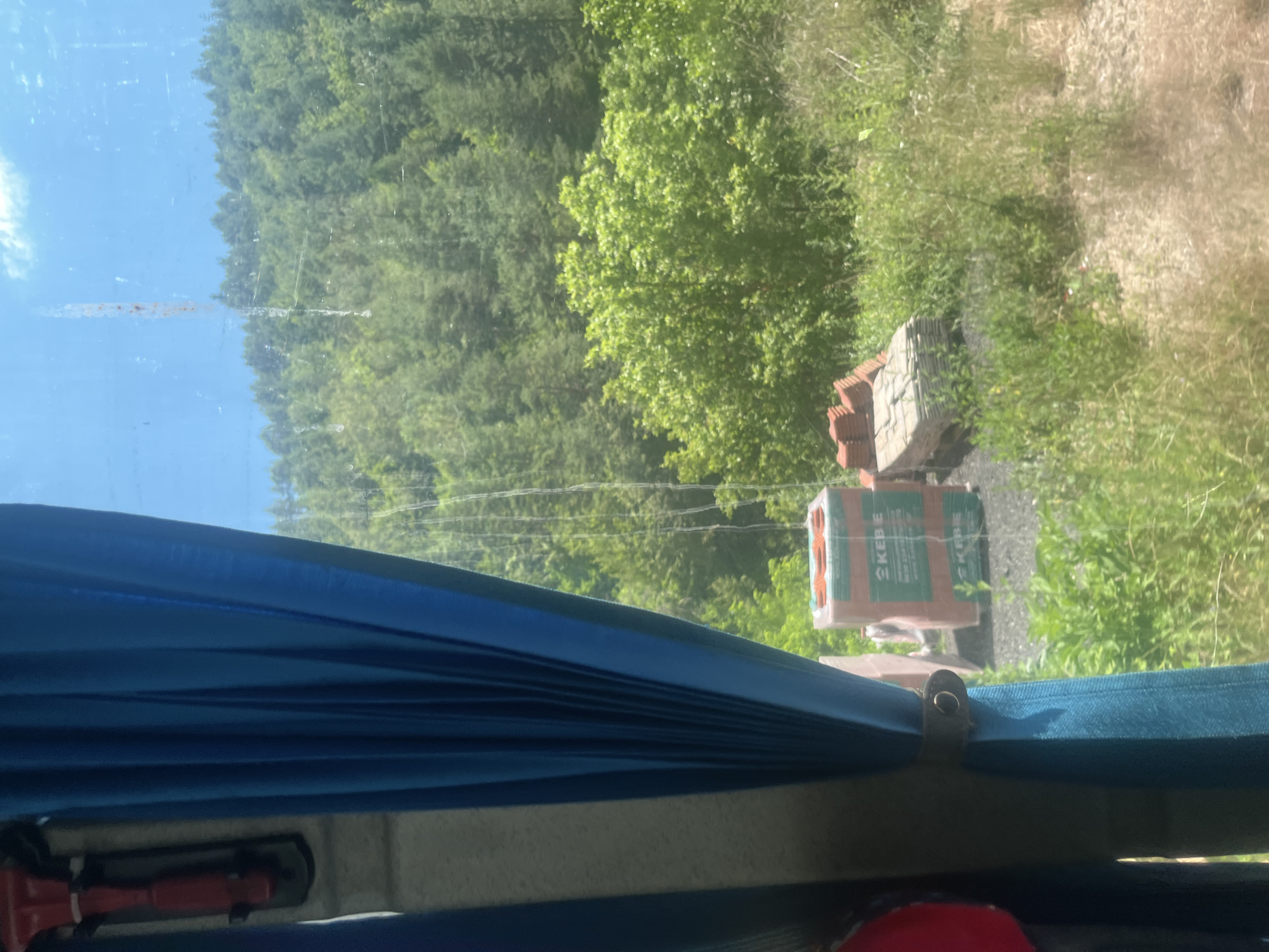 The image shows a view through a window, likely from a vehicle, looking out onto a wooded hillside under a blue sky. A blue curtain is partially drawn, obscuring the left side of the frame. Outside, on the ground, there are stacks of building materials, including what appear to be bricks and roof tiles. The vegetation is lush and green, suggesting a warm season. The scene is set in Nedelino, Bulgaria, a town known for its natural beauty and mountainous terrain.