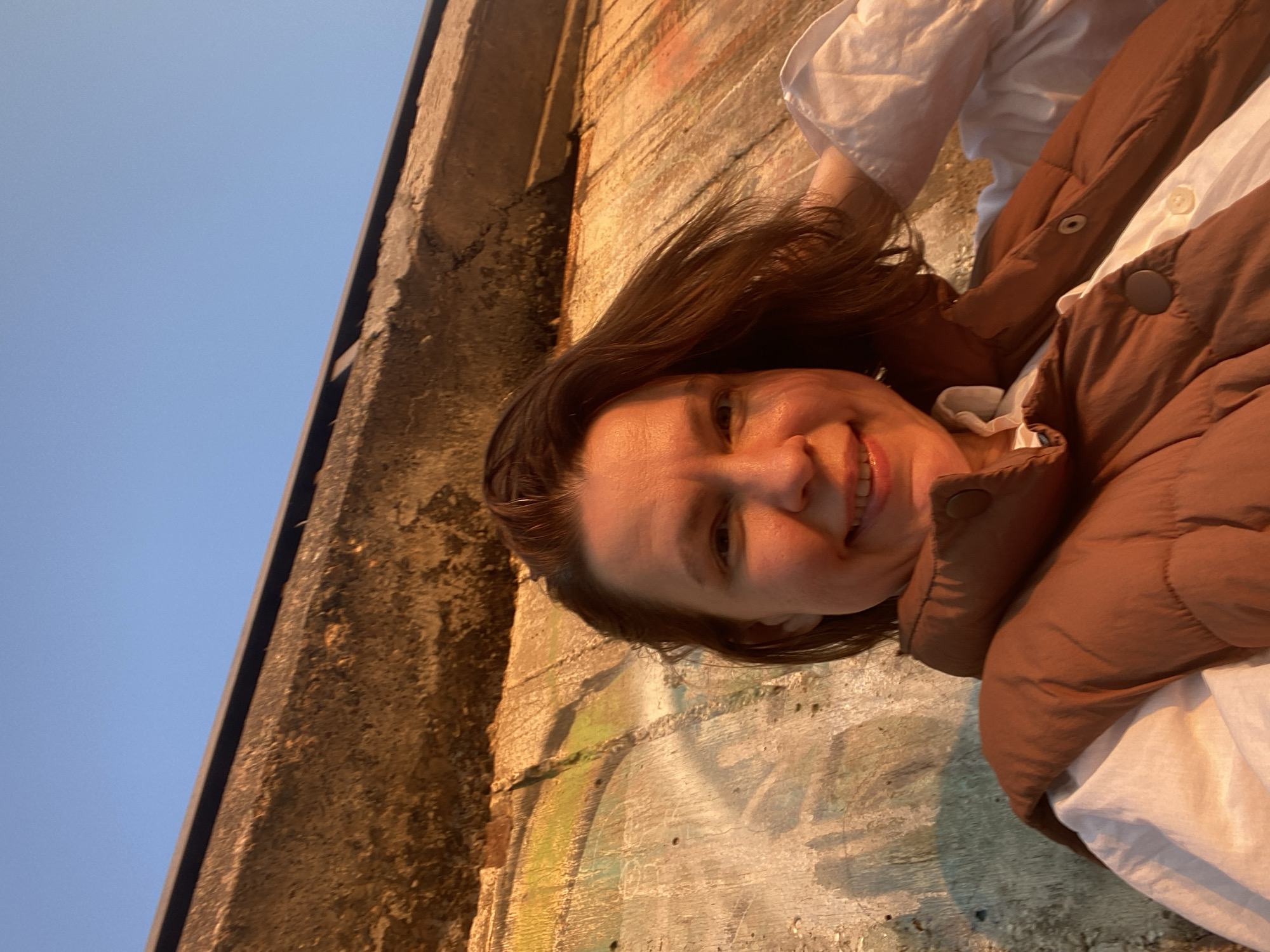 A woman with brown hair and a brown puffer vest over a white shirt is smiling at the camera. She is standing against a weathered, concrete wall that appears to have graffiti on it. In the upper left corner of the image is a clear blue sky. The photograph seems to be taken outside, possibly in the city of Chaikovsky, Russia, judging by the request for location context.