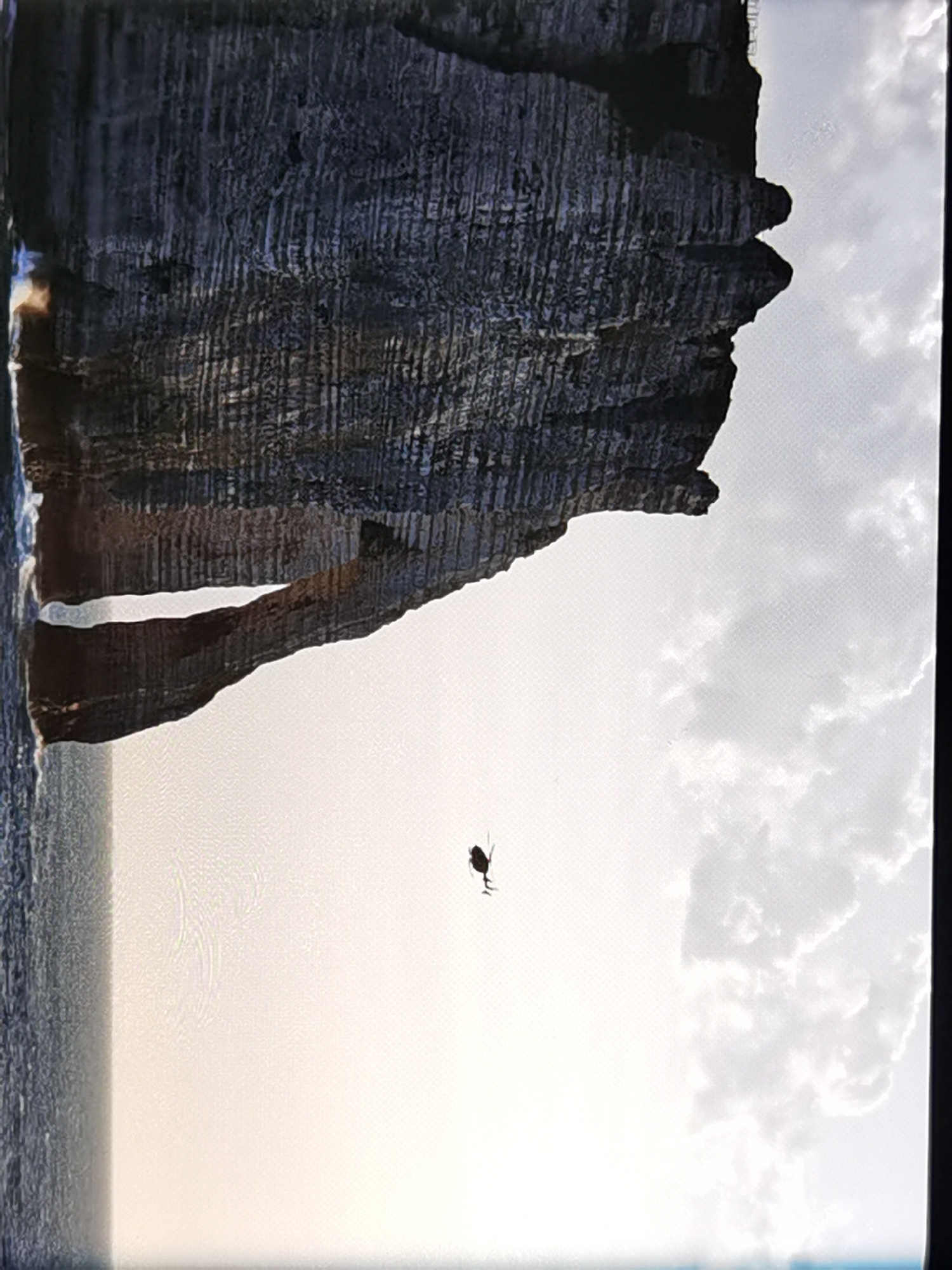 A helicopter is seen flying in the sky above the water, with a large rock formation behind it. The rock formation has a natural archway through it. The scene is likely Étretat, France, famous for its chalk cliffs. The image is taken from a low angle, looking up at the helicopter and cliffs. The sky is cloudy.