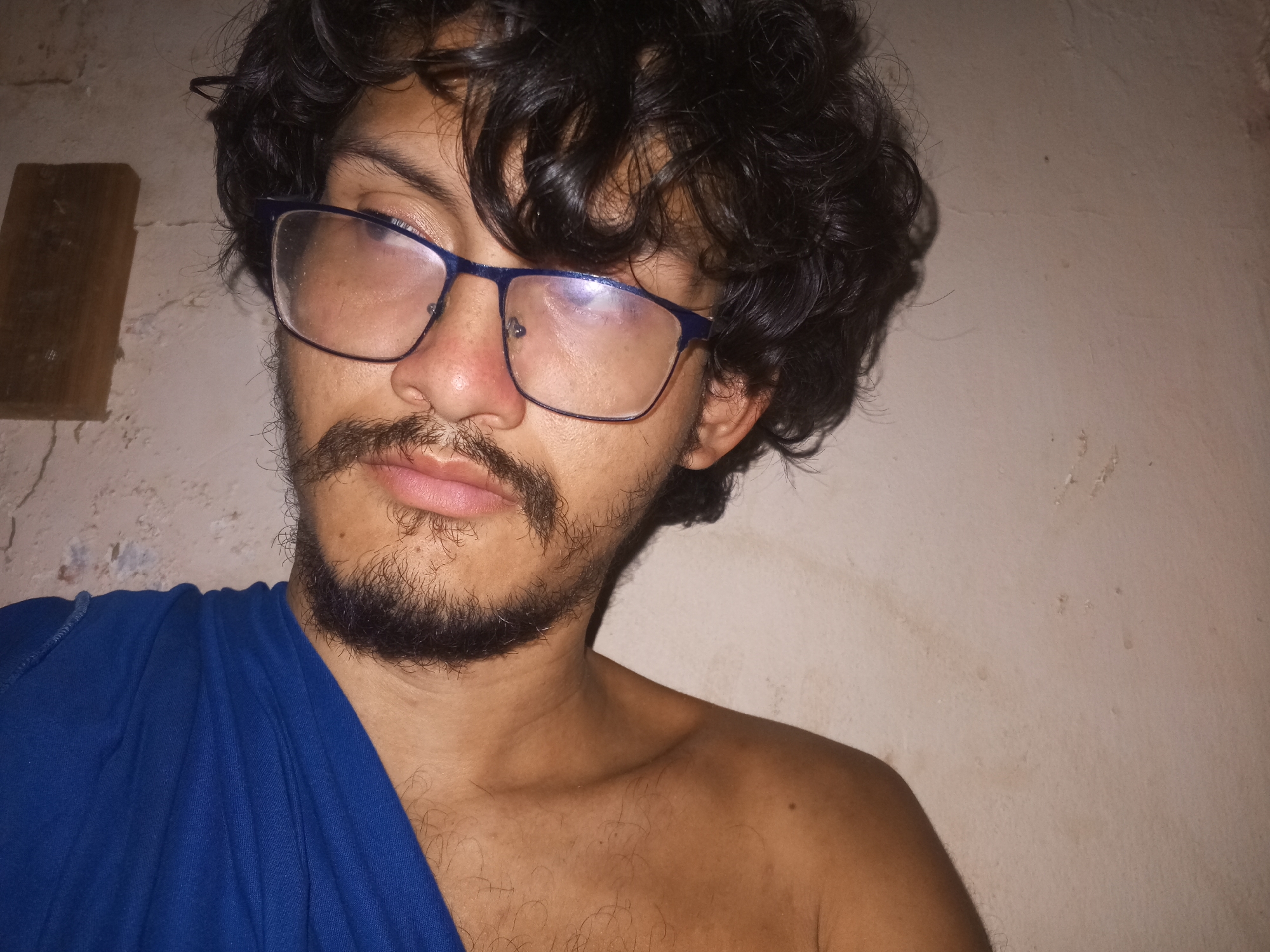 A close-up head and shoulder shot of a man with dark, curly hair and a beard, wearing glasses. He is wearing a blue shirt, partially draped over his shoulder. The background is a beige wall with some minor imperfections. The location is not discernible from the image.