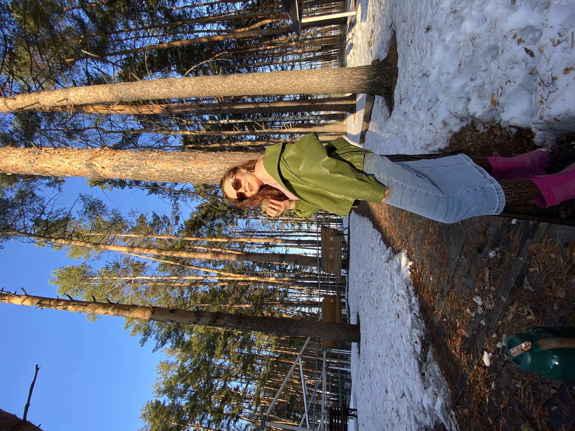 A woman sits on a wooden bench in a wooded area. She wears a green jacket, light blue jeans, and pink boots, with brown sunglasses on her face. The background features tall trees reaching towards a clear blue sky. Patches of snow are visible on the ground alongside the wooden path. The location is identifiable as the city of Chaikovsky, Russia. A metal fence and bench are partly visible further back in the scene.