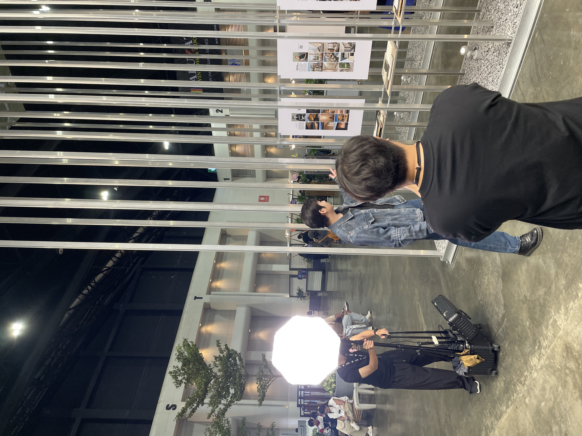 The image captures a scene indoors at an event in Pak Kret City Municipality, Thailand. Two people are looking at poster displays. One person wears a denim jacket and the other is in a black shirt. A photographer stands further away, operating a camera light and tripod. In the foreground, a gray floor leads to an area with seating and greenery, visible through the metal frame of the poster display. Overhead, an open ceiling structure with light fixtures is visible.