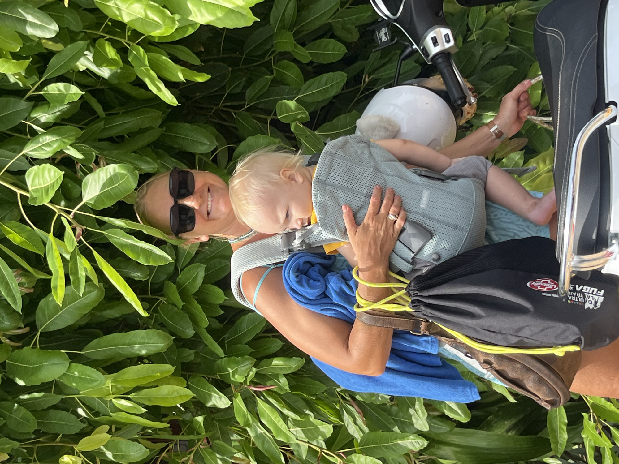 In the city of Mũi Né, Vietnam, a woman with blonde hair and sunglasses is standing in front of a dense green foliage backdrop. She is holding a baby in a carrier. The woman is wearing a blue top and has a blue towel draped over her arm. She is also carrying a brown leather bag and a black drawstring bag with yellow strings. Next to her, a white helmet rests on a black scooter. The scooter's handlebars and seat are visible. The woman's hand is reaching towards the scooter seat.