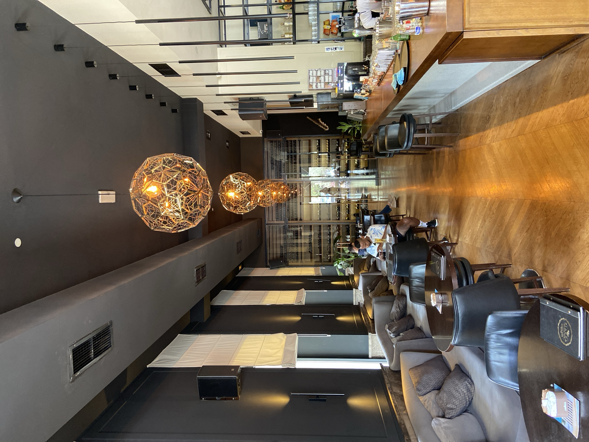 The image captures the interior of a modern and sophisticated bar or lounge, rotated vertically. The setting features a long wooden bar counter on the right side, stocked with various bottles, glassware, and shakers. A person in a white uniform is partially visible behind the bar, indicating active service.
On the left, a lounge area is furnished with a mix of comfortable grey upholstered sofas with patterned cushions and dark, possibly leather, chairs. These are arranged around dark, round tables. Several patrons are seated in this area; two men are visible towards the center-right, appearing relaxed, one looking at a phone or menu.
The decor is contemporary, characterized by dark grey walls and a warm wooden floor. Three prominent, ornate spherical pendant lights with a golden, geometric cage design hang from the ceiling, casting a warm glow. Additional linear track lighting and subtle wall sconces further illuminate the space. A large, glass-enclosed wine display spans a significant portion of the back wall, showcasing numerous bottles. Green plants are strategically placed, adding a natural element. White rectangular panels, possibly blinds or acoustic treatment, are visible along the lower section of the left wall. HVAC vents are integrated into the dark ceiling bulkheads.
The activity suggests a casual social gathering, with patrons relaxing and enjoying the ambiance, likely having drinks or light refreshments. The time of day appears to be daytime, given the presence of some natural light, although interior lighting is fully operational. A notable detail is the distinct "RIVA" logo visible on a black menu or binder on one of the tables in the foreground. This establishment is located in Budva, Montenegro.
