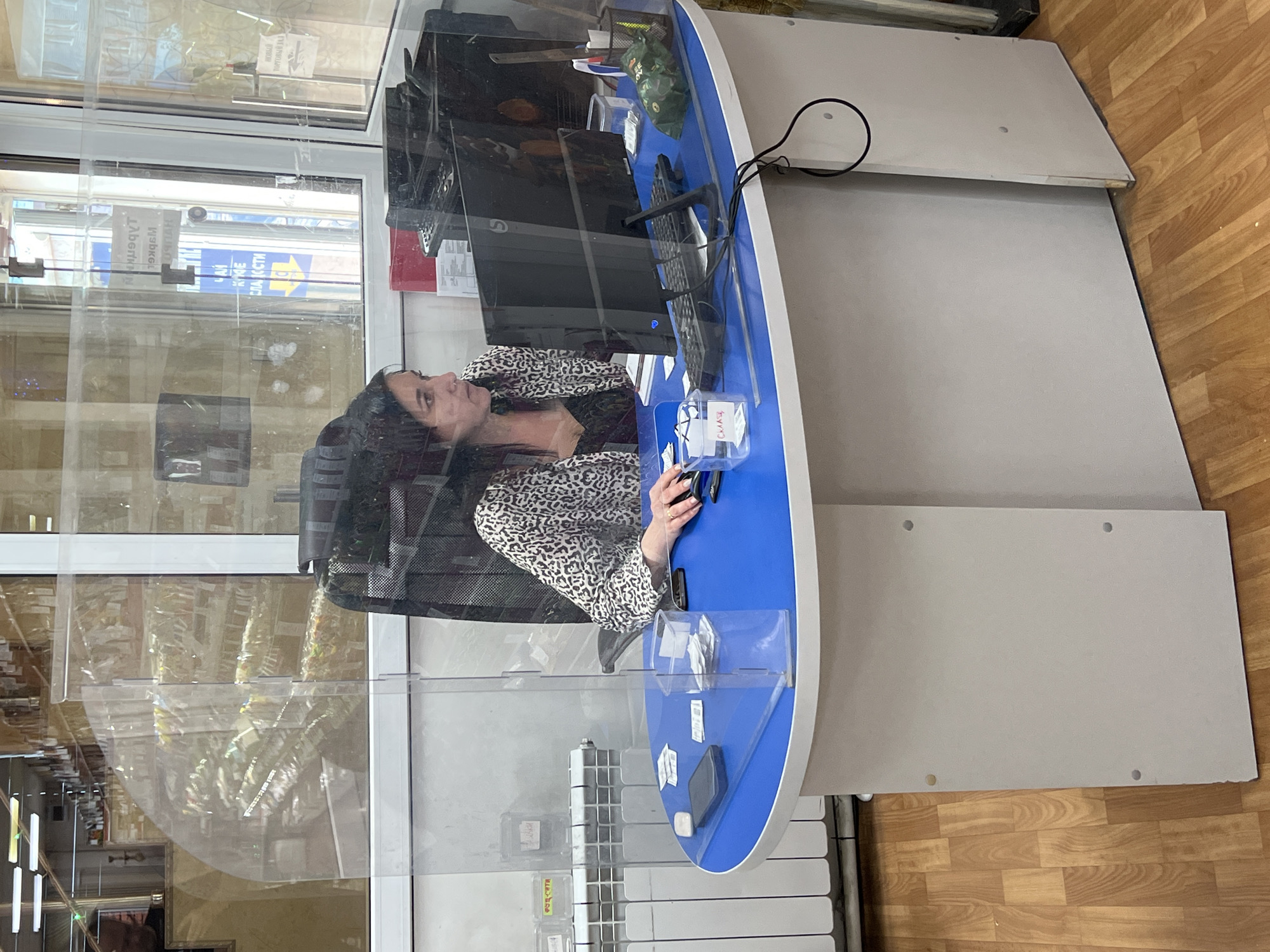 An indoor scene shows a woman seated at a blue-topped, curved reception desk, positioned behind a transparent protective barrier. The woman, with long dark hair and wearing a patterned jacket, is looking towards her right, with her hands resting near a computer keyboard and mouse, indicating she is working. A black computer monitor is centered on the desk, alongside other office items like a pen holder and clear plastic boxes. One clear box on the desk is labeled "Склад" (Russian for "Warehouse" or "Stock").
The transparent barrier creates reflections of the surrounding environment. These reflections show snippets of Russian text from signs, including "РУБЛИ" (Rubles), "ОБМЕН ВАЛЮТ" (Currency Exchange), "ПОРТОВЫЙ ГРУЗ" (Port Cargo/Harbor Freight), and "НАШИ УСЛУГИ" (Our Services), along with a yellow arrow. These cues suggest a financial institution or a currency exchange office in Krasnodar, Russia.
The desk features a light grey base, and the floor is covered with wooden planks. The lighting appears to be bright, likely from overhead lights and possibly natural light entering through windows, as suggested by the reflections. The woman's expression is neutral and focused on her task. The overall impression is that of a customer service or administrative workspace during daylight hours.