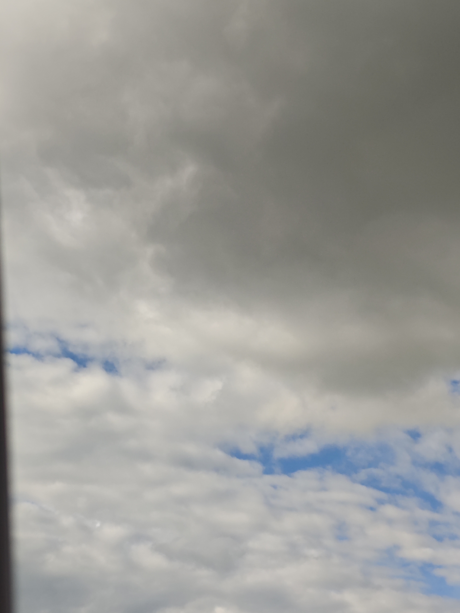 The image displays a sky with varied cloud formations. The upper portion of the image is dominated by dark, heavy clouds, suggesting overcast conditions. Towards the middle and lower sections, lighter, more diffused clouds are visible, interspersed with patches of blue sky. The lighting indicates it is daytime. There are no discernible landmarks or specific features that would allow for identification of a location within Gubithsky Selsovet, Belarus. The image does not contain any people, subjects, objects, or actions.