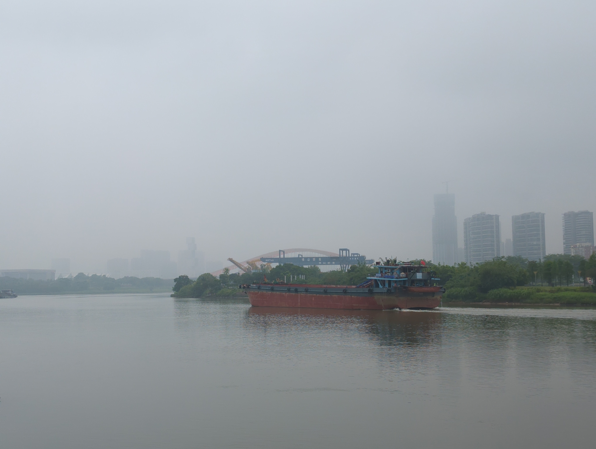 A cargo ship moves across a wide body of water. The ship is primarily red with a blue and gray upper structure. A faint wake trails behind the vessel.
The background includes an orange-colored bridge with blue gantry cranes. Further in the distance, buildings are visible, including high-rise structures. The location is Shunde District, China.
The overall atmosphere is hazy, obscuring details in the background. The water is relatively calm, reflecting the overcast sky. A tree-lined bank runs along the right side of the river.