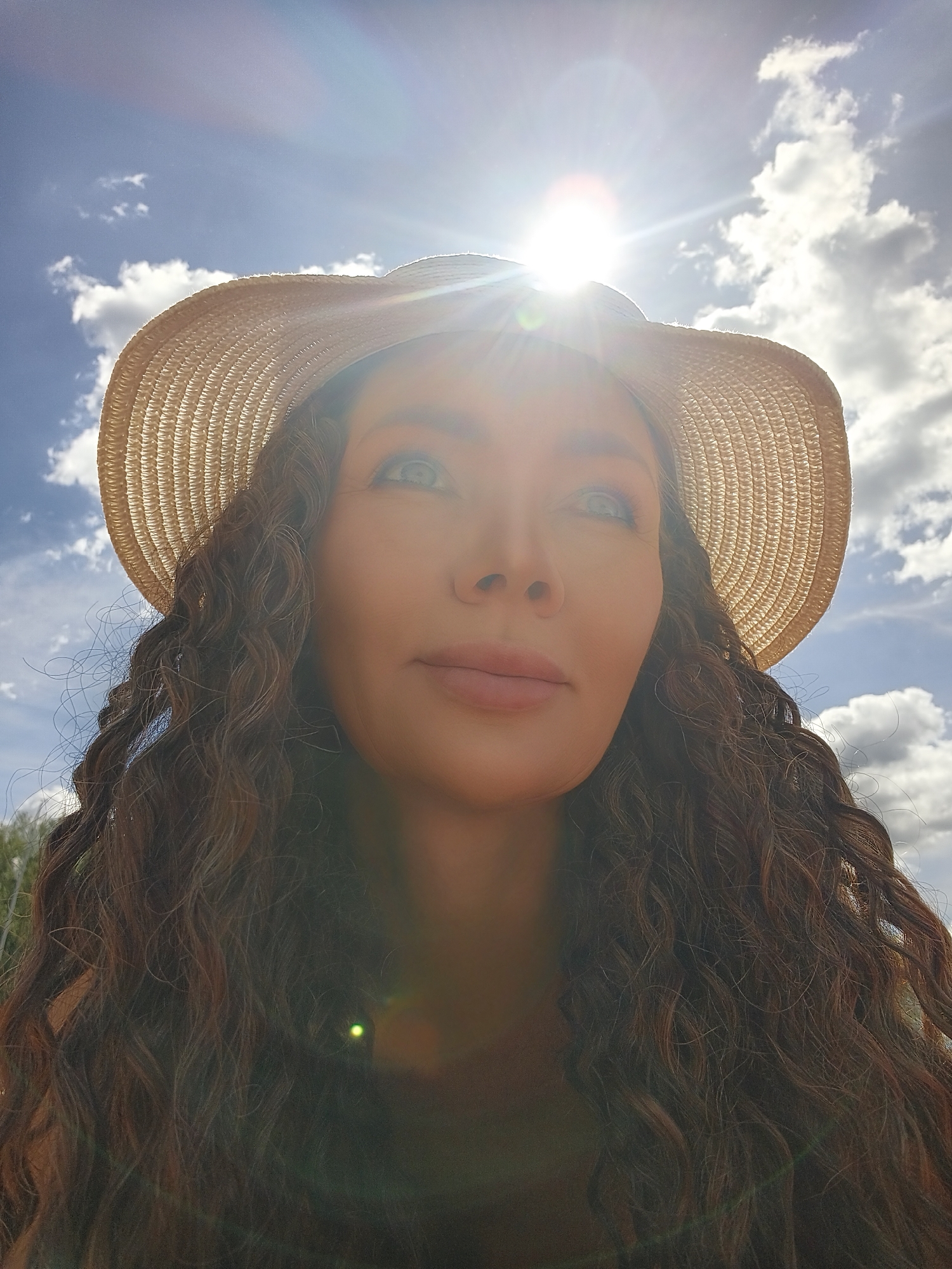 A low-angle, close-up shot captures a woman from the chest up, looking up towards the sun, which is directly overhead and heavily flares the lens. She is wearing a wide-brimmed straw hat, casting a shadow over the top of her head. Her long, dark, curly hair is visible on either side of her face and shoulders. The background is a bright blue sky with scattered white clouds, suggesting a sunny day. The sunlight is intense, creating lens flares and glares that obscure parts of the image, particularly the top of her face and the hat. Her expression is serene, with her eyes gazing upward. The overall impression is one of warmth and brightness, possibly a summer day outdoors. There is no visible text.