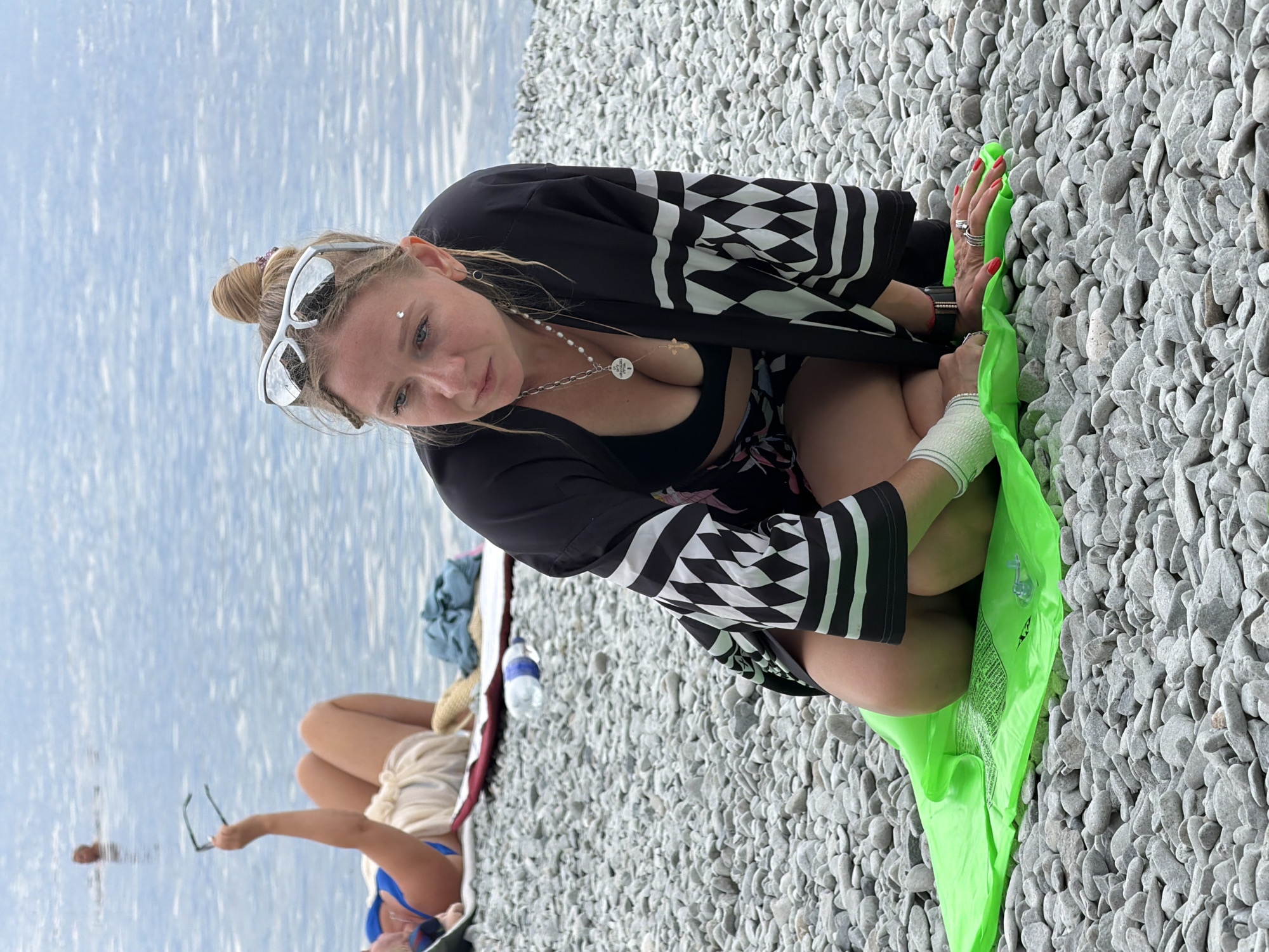 A woman with blonde hair styled in a bun and wearing sunglasses on her head, a black and white patterned kimono over a black bikini top and patterned shorts, is kneeling on a bright green inflatable mat on a pebble beach. She has a silver necklace with a cross pendant and a circular charm. Her hands, adorned with rings and red nail polish, are resting on the mat. To the left, another person is lying down on a red mat, holding up a pair of eyeglasses, wearing a blue bikini top and light-colored shorts. In the background, the calm, rippling surface of the sea is visible. A person can be seen in the water in the distance. The location is the pebble beach of Partenit, Ukraine.