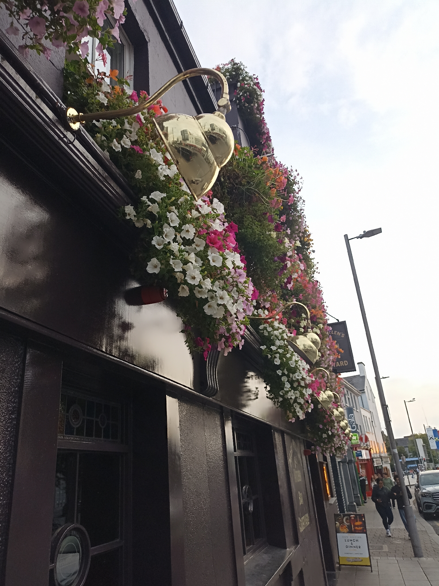 The image captures a low-angle, wide shot of the exterior of a building in Galway, Ireland. The building is dark brown and adorned with an abundance of colorful flowers cascading from window boxes. Golden heart-shaped lamps are affixed to the building, casting a warm glow. A sign above the entrance reads "St. Helen's" and "Bernard". To the right, a street scene unfolds with a lamppost, a sidewalk, and several people walking. A car is visible on the street. The sky is overcast. The overall impression is of a vibrant and inviting establishment in a bustling urban environment.