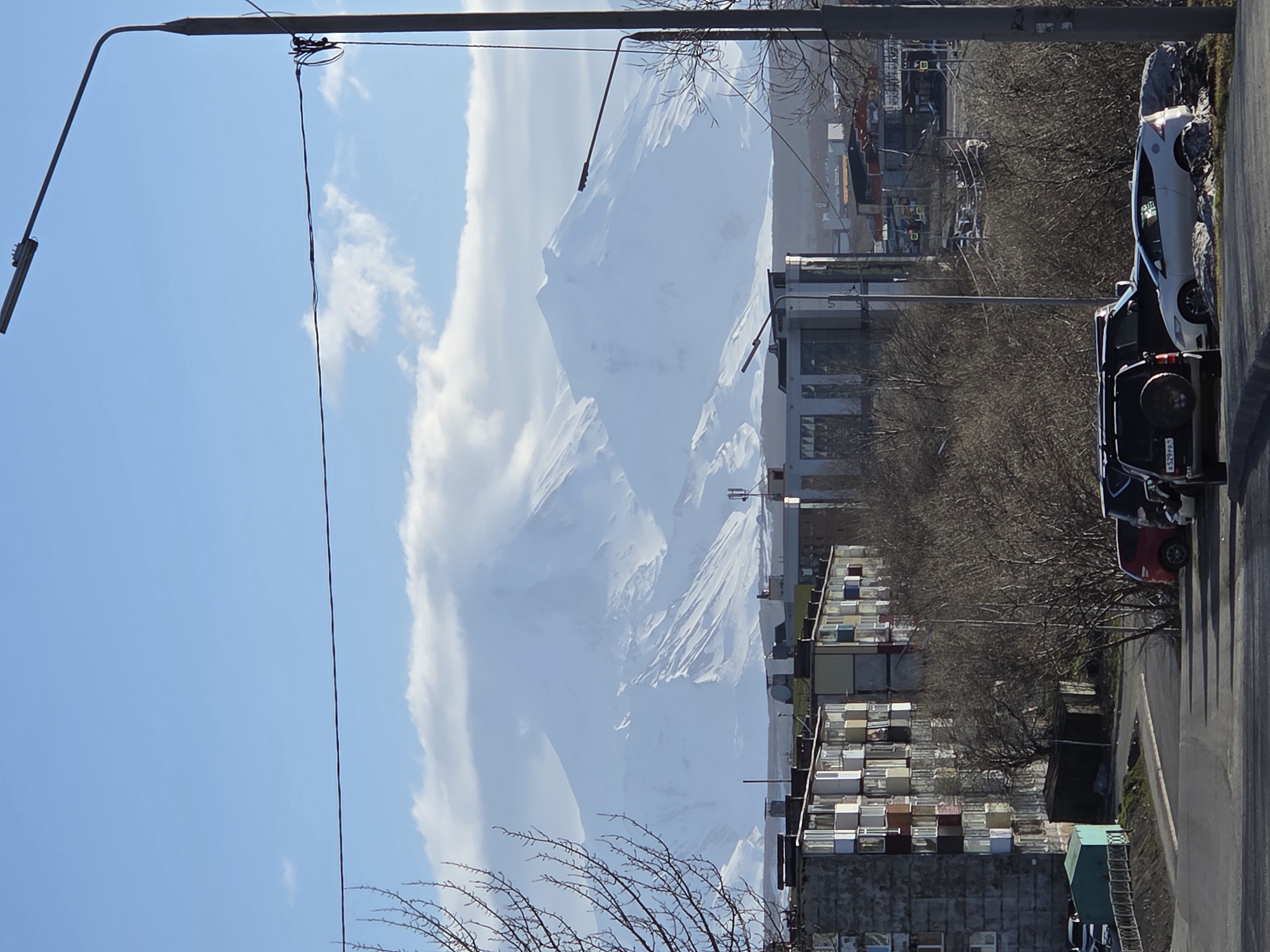 The image depicts a street scene in a city, featuring a mountain backdrop partially obscured by clouds. A street runs along the right side of the image, with several parked cars. Along the street, there's a mix of buildings, including what appears to be an apartment complex or dormitory-style structure with numerous windows, as well as some industrial or commercial buildings. Bare trees and utility poles line the street. The sky is blue with scattered clouds, and the distant mountain is snow-covered.