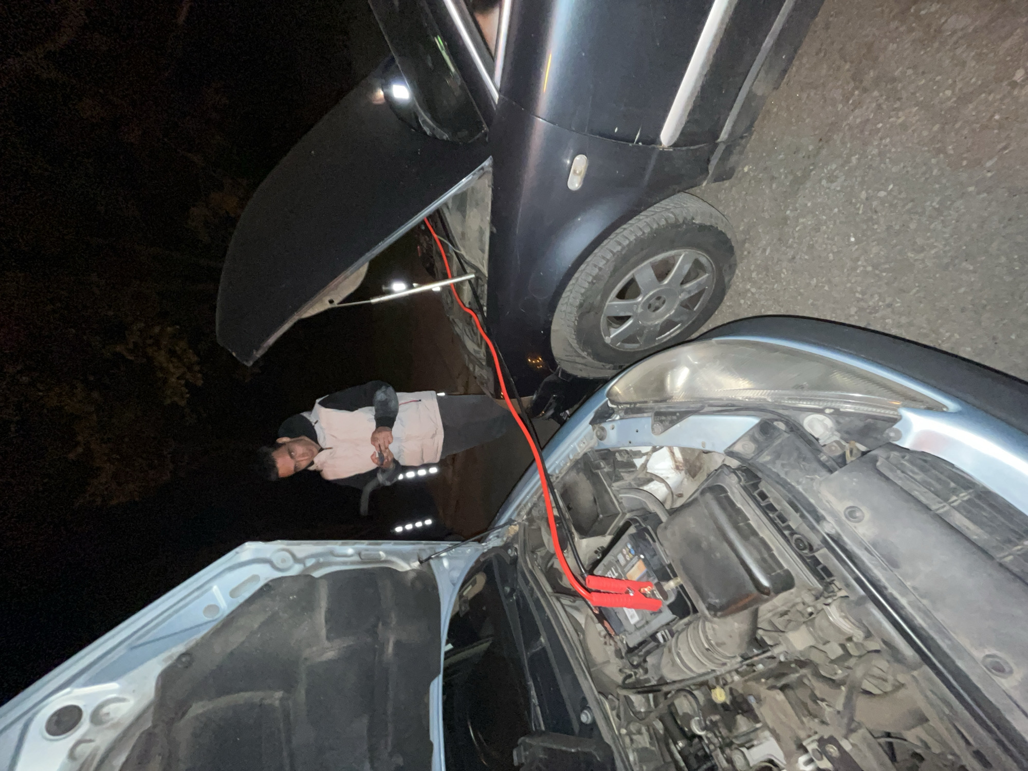 The image, captured at night in Sofia, Bulgaria, depicts a car being jump-started. In the foreground, a light blue or silver car has its hood fully open, revealing the engine bay. A red jumper cable is prominently connected to the positive terminal of the car's battery, which has the brand name "VARTA" visible on its label. Various engine components, including the air filter housing and associated hoses, are also visible.
A person, appearing to be a man, is positioned in the mid-ground, leaning over and looking down into the engine bay of the light blue car. He is wearing a light-colored top with dark sleeves. His presence indicates active involvement in the jump-starting process.
Behind the light blue car, a second, darker colored vehicle (likely black or dark blue) is parked. The red jumper cable extends from the foreground car's battery towards this darker vehicle, suggesting it is either providing power to or receiving power from it. The side of the darker car, including one of its wheels, is visible.
The scene takes place outdoors on an asphalt surface, illuminated by artificial lights reflecting off the car bodies. The dark background confirms the nighttime setting. The overall activity centers around practical car maintenance, addressing a potential dead battery.