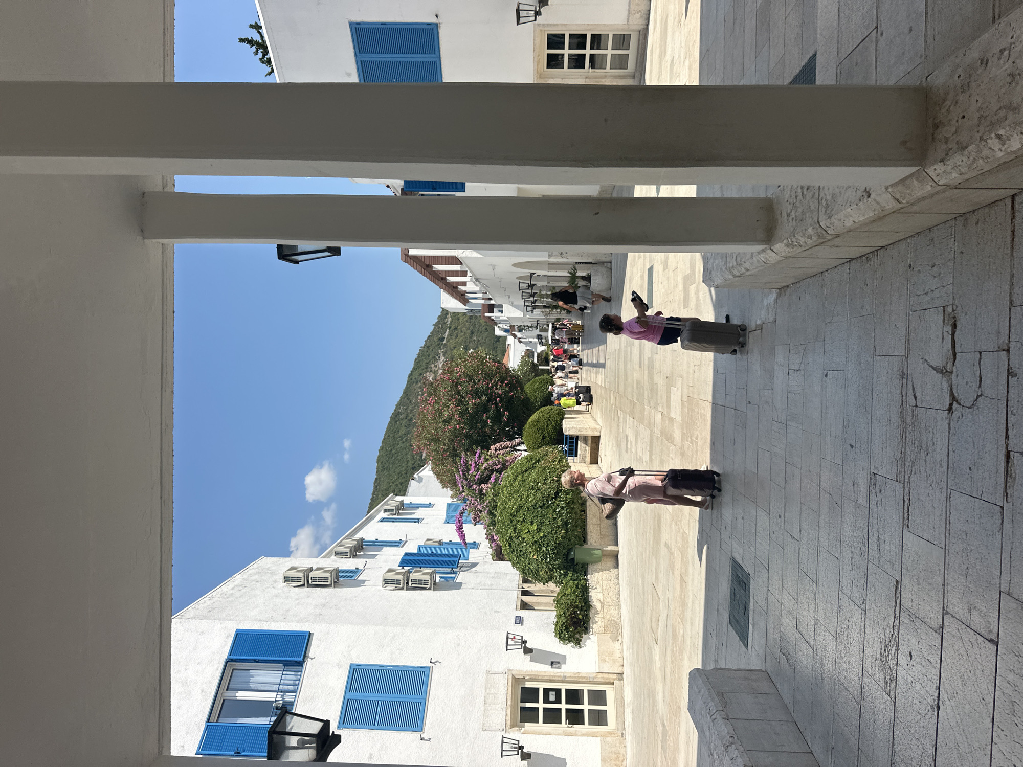 The image depicts a sun-drenched, pedestrianized street in the city of Budva, Montenegro, likely within a resort or historic area. The architecture is characterized by white buildings with vibrant blue shutters and doorways, set against a backdrop of lush green hills under a clear blue sky. The street is paved with large, light-colored stone tiles, with architectural elements like white pillars and decorative stonework framing the scene.
In the foreground, two women are visible. One, on the left, is a Caucasian woman with blonde hair, wearing a pink outfit and pulling a dark suitcase. She is walking away from the camera. To her right and slightly further down the street, a younger Caucasian girl in a pink shirt and dark shorts is also pulling a suitcase, walking towards the right. Several other individuals and groups are scattered along the street, some with luggage, suggesting a travel or tourist context.
The buildings feature rows of air conditioning units mounted on their facades, and outdoor lighting fixtures are visible along the covered walkways. Planters with greenery and flowering bushes, including a vibrant pink flowering tree, add color and texture to the environment. The overall impression is one of a well-maintained, picturesque European town on a bright, sunny day.
