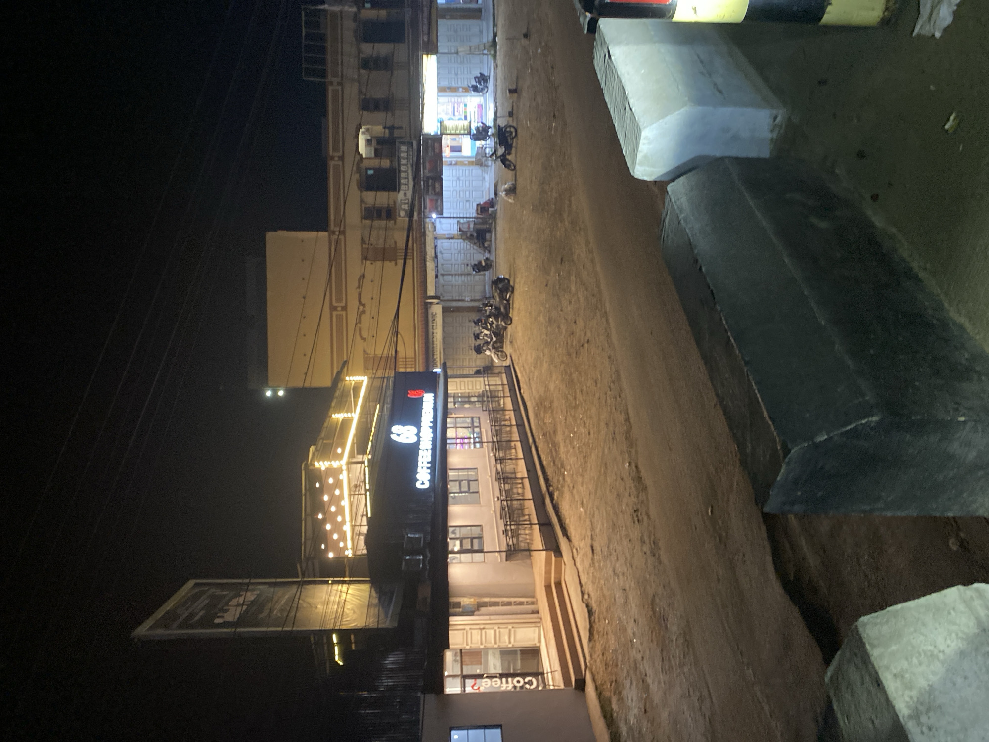 The image depicts an outdoor street scene at night in Seuneubok Teungoh, Indonesia, illuminated by artificial lights. The sky is dark, indicating the time of day.
On the left side of the frame, a row of multi-story buildings lines the street. A prominent building features a large, brightly lit sign that reads "COFFEE HOPPERIAN" in white and red lettering. Above this sign, an area is adorned with string lights, creating an inviting glow. Below "COFFEE HOPPERIAN," another sign or window displays "Coffee?". Further down the street, another building has a sign with the text "AL-BAROKAH," and partially visible below it is the word "CLINIC." Overhead, multiple utility lines are visible against the dark night sky.
The ground is an unpaved or dirt road that recedes into the distance. Several motorcycles are parked along this road, suggesting activity within the buildings. On the right side of the frame, concrete blocks or barriers line the edge of the road, and a yellow and black striped barrier is partially visible in the upper right.
The scene portrays a commercial or semi-commercial area during the evening, with businesses open and their signs lit. There is no specific activity or event captured, but the parked vehicles imply patronage of the establishments. The overall atmosphere is one of a quiet but active street at night, typical of an urban or semi-urban setting.