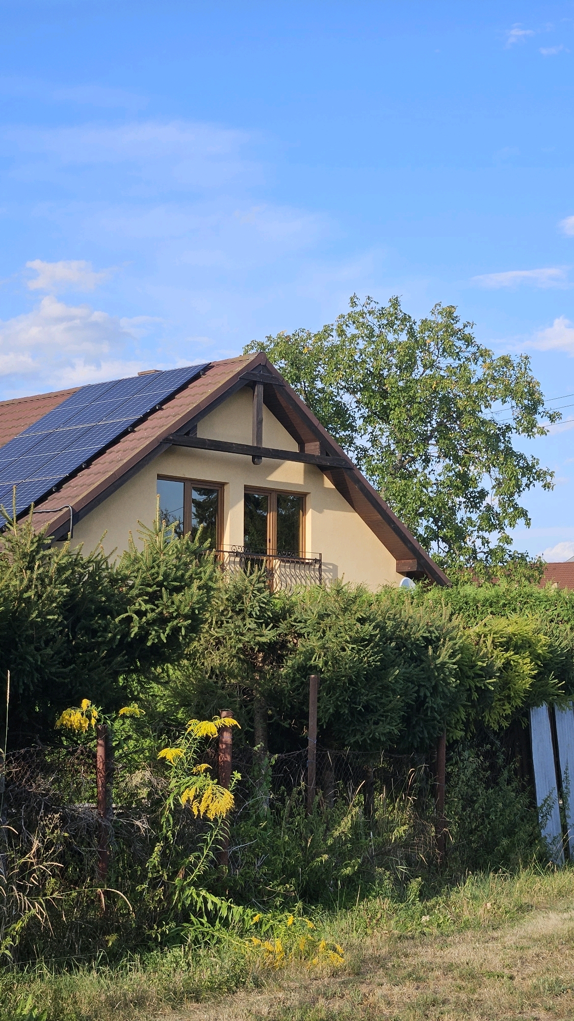This is a vertical, eye-level, medium shot of a house in Prešov, Slovakia, with solar panels on the roof. The house is light yellow with a brown roof and wooden-framed windows. A balcony with a wrought-iron railing is visible above a dense hedge of evergreen trees. A large, leafy tree is behind the house. The foreground features a wire fence with rusty metal posts and patches of yellow wildflowers and green grass. The sky is blue with a few white clouds.
