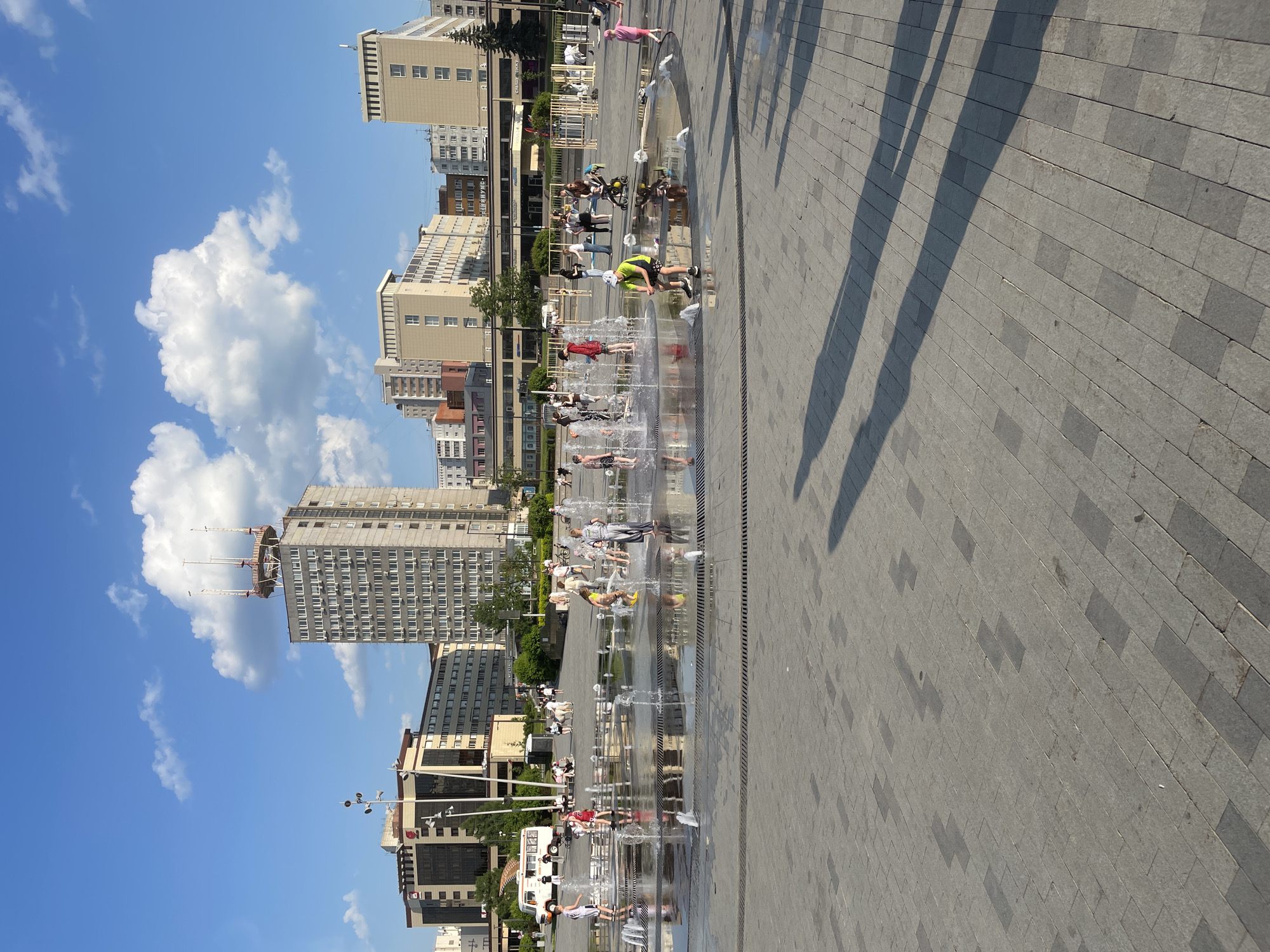 The image depicts a city square in Perm, Russia, with a fountain as its central feature. Numerous individuals, including children and adults, are playing in and around the water jets of the fountain. The square is paved with gray stone blocks. Shadows cast across the square suggest a sunny day. The urban backdrop includes various buildings of different heights and architectural styles, with a mix of residential and commercial properties. A clear blue sky with white clouds is visible overhead.