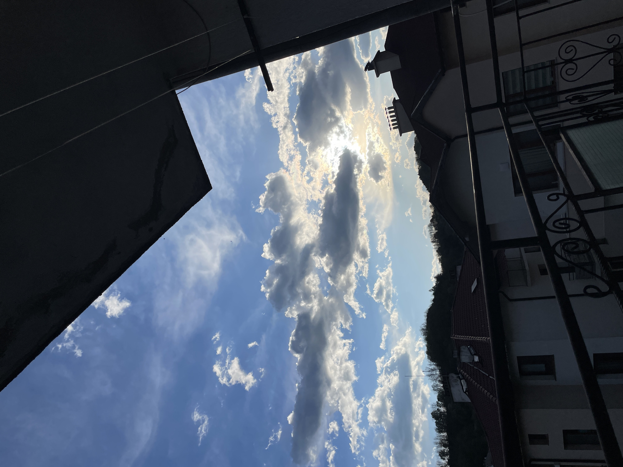 The image shows a view of the sky with numerous white clouds, partially obscuring the sun. The view is framed by architectural elements, including a dark gray, angled structure on the left and a building with a red-tiled roof and ornate ironwork on the right. In the distance, a hillside with trees is visible. The photo was taken in Zlatograd, Bulgaria.