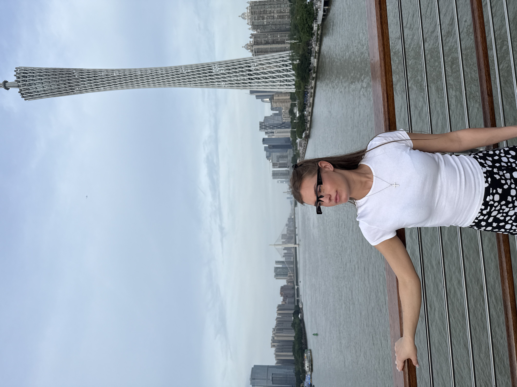 A full-body outdoor shot shows a young Caucasian woman with long brown hair tied back with black ribbons, wearing a white t-shirt and a black and white polka dot skirt. She is standing on a bridge, resting her left arm on a wooden railing. She is wearing black-rimmed glasses. In the background, a river flows through a city skyline featuring numerous skyscrapers and a distinctive, latticed white tower. The sky is overcast. The image appears to be taken in Guangzhou, Yuexiu District, China, given the prominent Canton Tower in the background.
