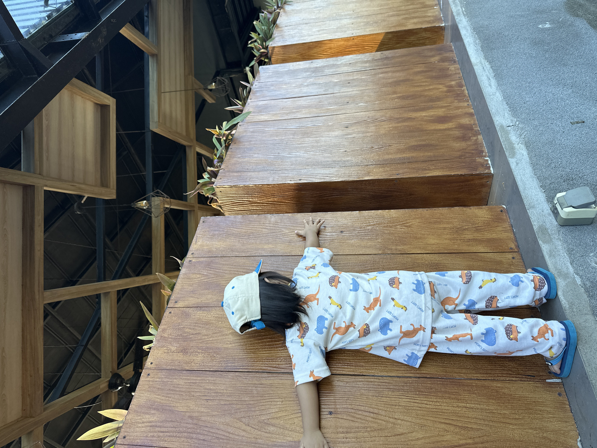 A child wearing a light-colored pajama set patterned with animals, a blue baseball cap, and blue sandals lies on a set of wooden stairs, appearing to mimic a "starfish" position with arms and legs outstretched. The stairs have plants growing along their edge, suggesting an outdoor or partially outdoor setting. To the left, a complex structure of wooden beams and metal supports can be seen, possibly part of a building’s architectural design. To the right, the steps end against a concrete walkway, with an electrical outlet visible. The location is in Unknown, Indonesia.