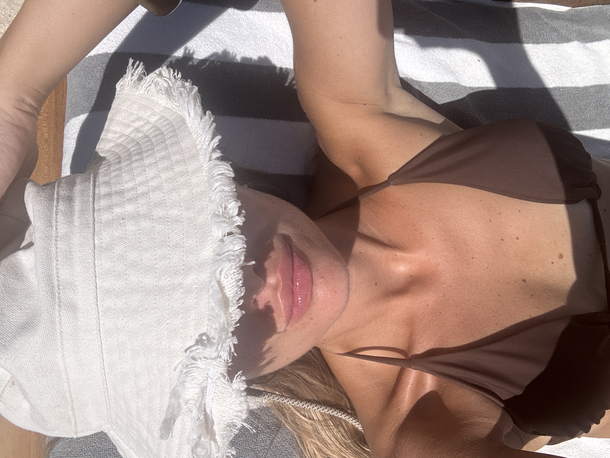 A person lies on a striped towel outdoors, wearing a brown bikini top and a white hat with a frayed edge. The location is presumably in the city of Unknown, Indonesia. The person's face is partially obscured by the hat. The scene is illuminated by strong sunlight, creating shadows. The person's skin appears tanned.