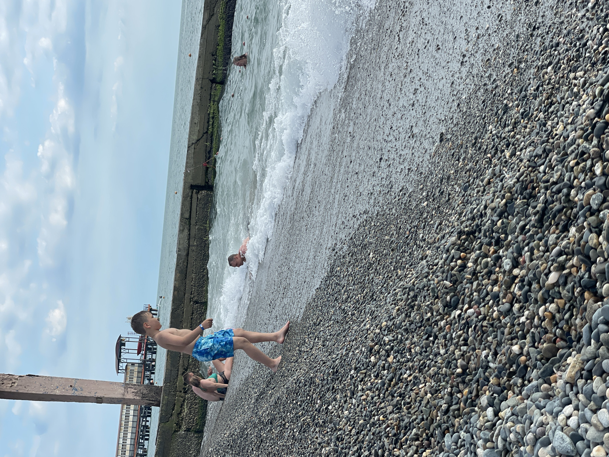 This media file depicts a sunny day at a pebble beach in Sochi, Russia. The scene is oriented vertically, with the sky occupying the top portion and the beach and sea filling the lower part.
Several individuals are visible. In the foreground, a young boy is standing on the pebble shore, wearing blue patterned swim trunks. He is looking towards the sea. Beside him, a person is sitting on the pebbles, wearing a green bikini. Further back in the water, a man is partially submerged and appears to be swimming or playing in the waves. The sea is choppy, with white foam from breaking waves crashing onto the shore.
In the background, a concrete pier runs along the water's edge, partially covered in green algae. Beyond the pier, structures of a building or resort are visible, suggesting a developed coastal area. The sky is partly cloudy, with white clouds scattered across a light blue expanse.
The overall atmosphere is one of leisure and outdoor recreation on a warm day. The presence of people enjoying the water and the sun indicates it is likely midday or afternoon during the warmer months. The pebble beach and the characteristic green algae on the pier are consistent with the coastal environment of Sochi. No visible text is present in the image.