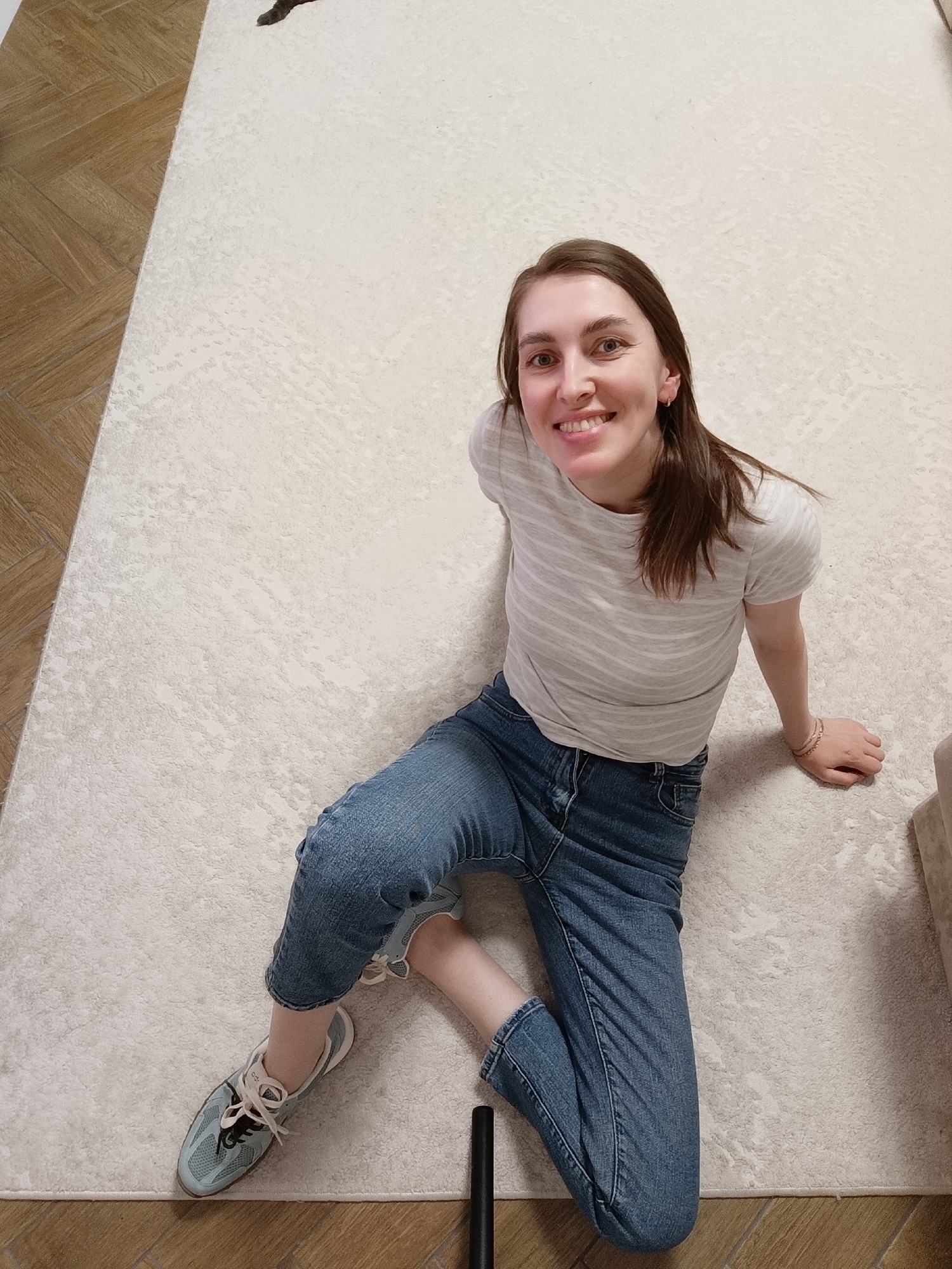 A young woman is captured from a high-angle perspective, sitting on a light-colored, textured rug indoors. She has brown hair and is smiling broadly, looking directly at the camera. She is wearing a light grey and white striped short-sleeved t-shirt, blue jeans that appear cropped or rolled up, and light blue sneakers with white laces. Small hoop earrings are visible, along with multiple delicate bracelets on her right wrist.
The rug covers most of the visible floor area. To the left, a section of wood-look tiled flooring, arranged in a herringbone pattern, is visible. A portion of a light-colored piece of furniture, possibly a sofa, can be seen on the right side of the image. A black cylindrical object is situated near her left foot on the rug. The setting appears to be a casual indoor space, possibly a living room, with bright, natural-looking illumination. The woman's expression conveys happiness and a relaxed demeanor. There is no visible text.