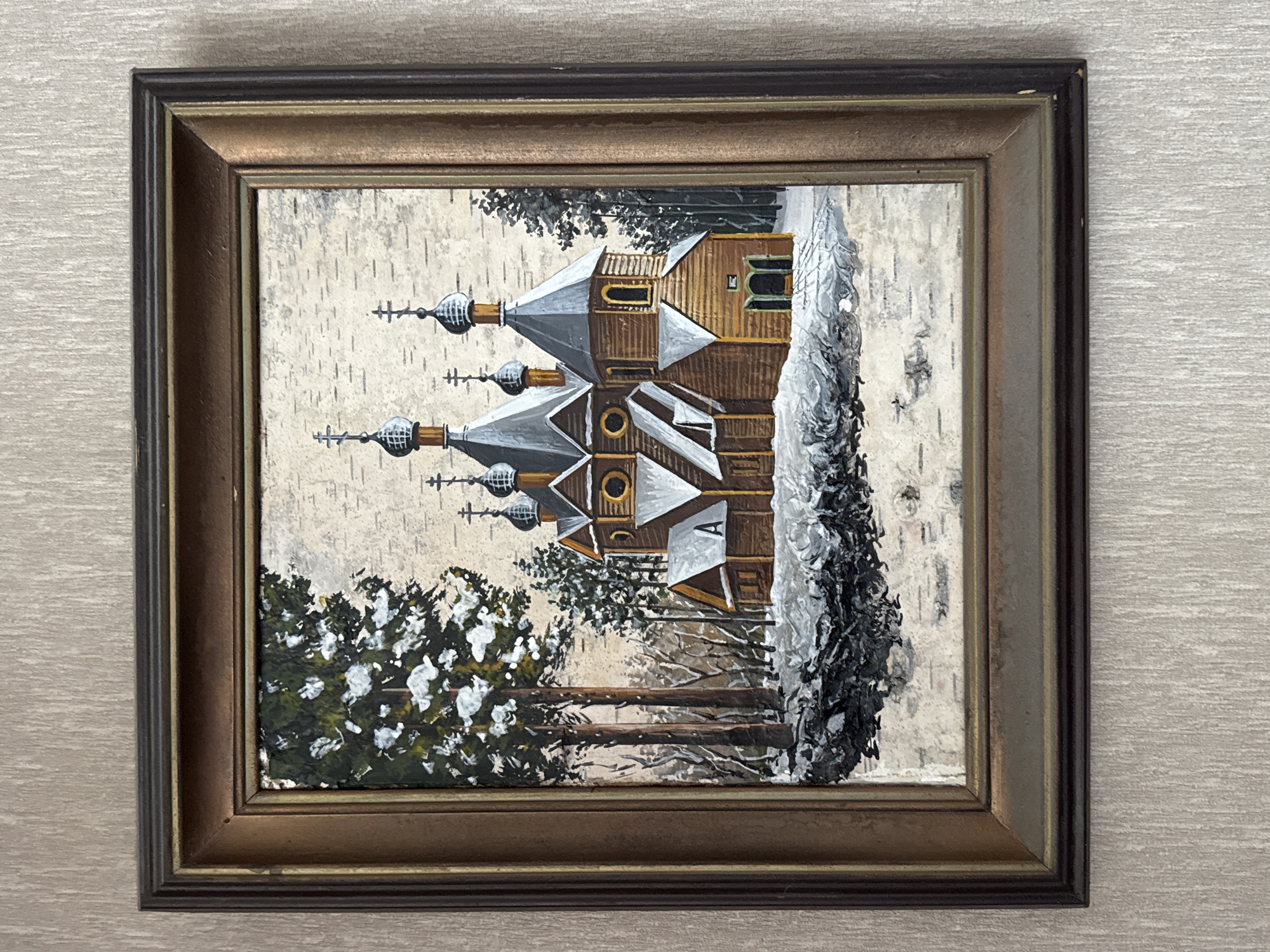 A framed painting depicts a wooden church with multiple onion domes covered in snow, set amidst a winter landscape with snow-covered trees. The painting appears to be rendered on birch bark, indicated by the textured, pale background. The church is predominantly brown with grey roofs and decorative elements. Snow is visible on the ground and on the church's structures. In the foreground, there's a textured representation of snow and possibly branches. The painting is housed in a multi-layered wooden frame with a metallic sheen. The background wallpaper is textured and light beige. This image does not depict any specific location in Bishkek City, Kyrgyzstan.