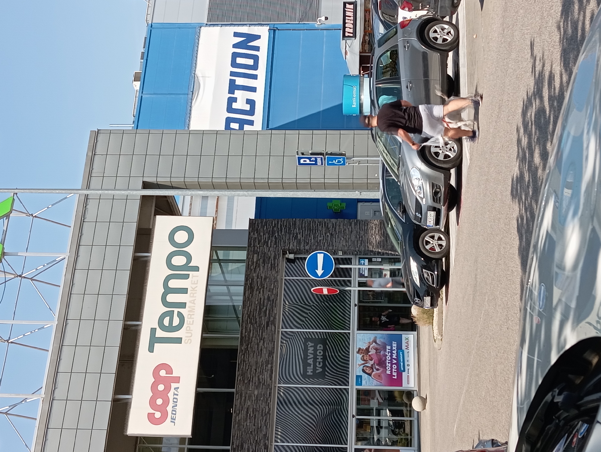 The image shows the entrance to a Coop Jednota Tempo supermarket in Nitra, Slovakia. A man is walking a dog in front of the supermarket. The supermarket has a large sign with the Coop Jednota logo and "Tempo SUPERMARKET" written on it. There are several cars parked in front of the supermarket. The weather is sunny.