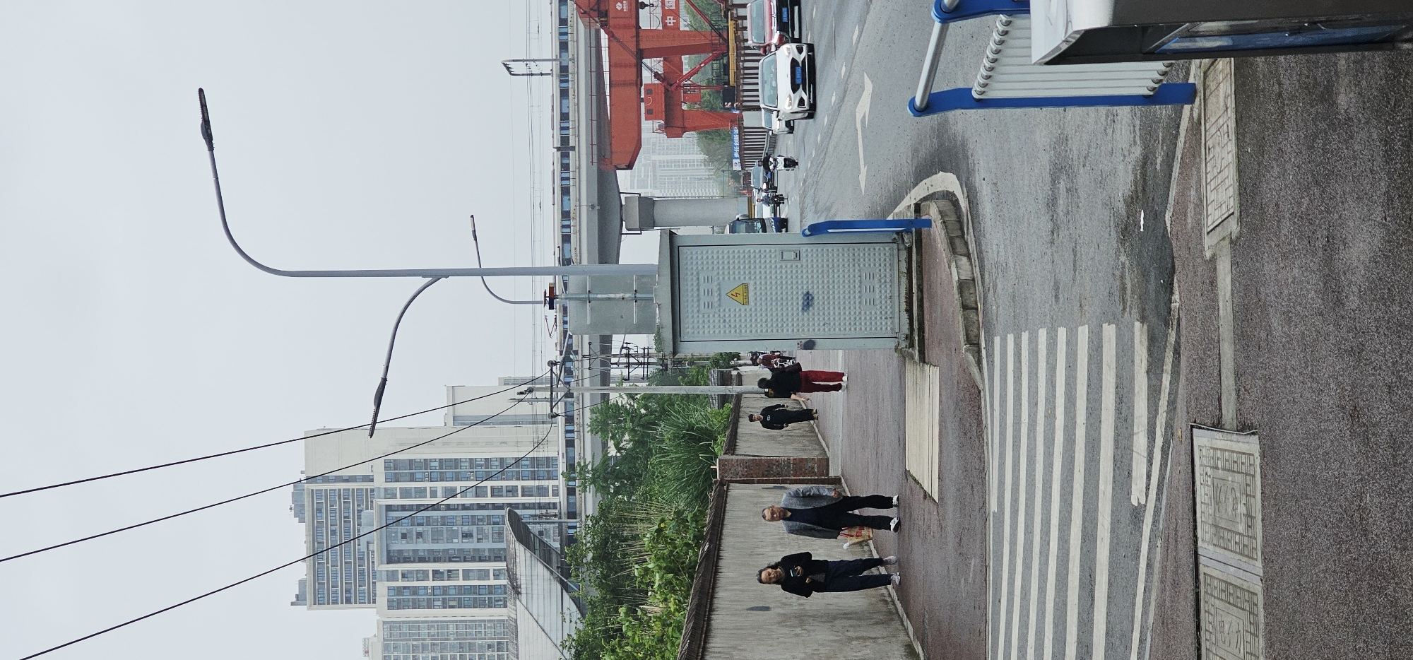 The image captures a street scene in the Chongqing High-tech Zone, China. The perspective is tilted, showing a sidewalk on the right and a road on the left. Several people are walking on the sidewalk, one pair is closer to the camera. A utility box is placed near the crosswalk. The road has pedestrian markings. In the background are buildings, an overpass structure, and a red crane. Streetlights and power lines stretch across the sky. The sky appears overcast.